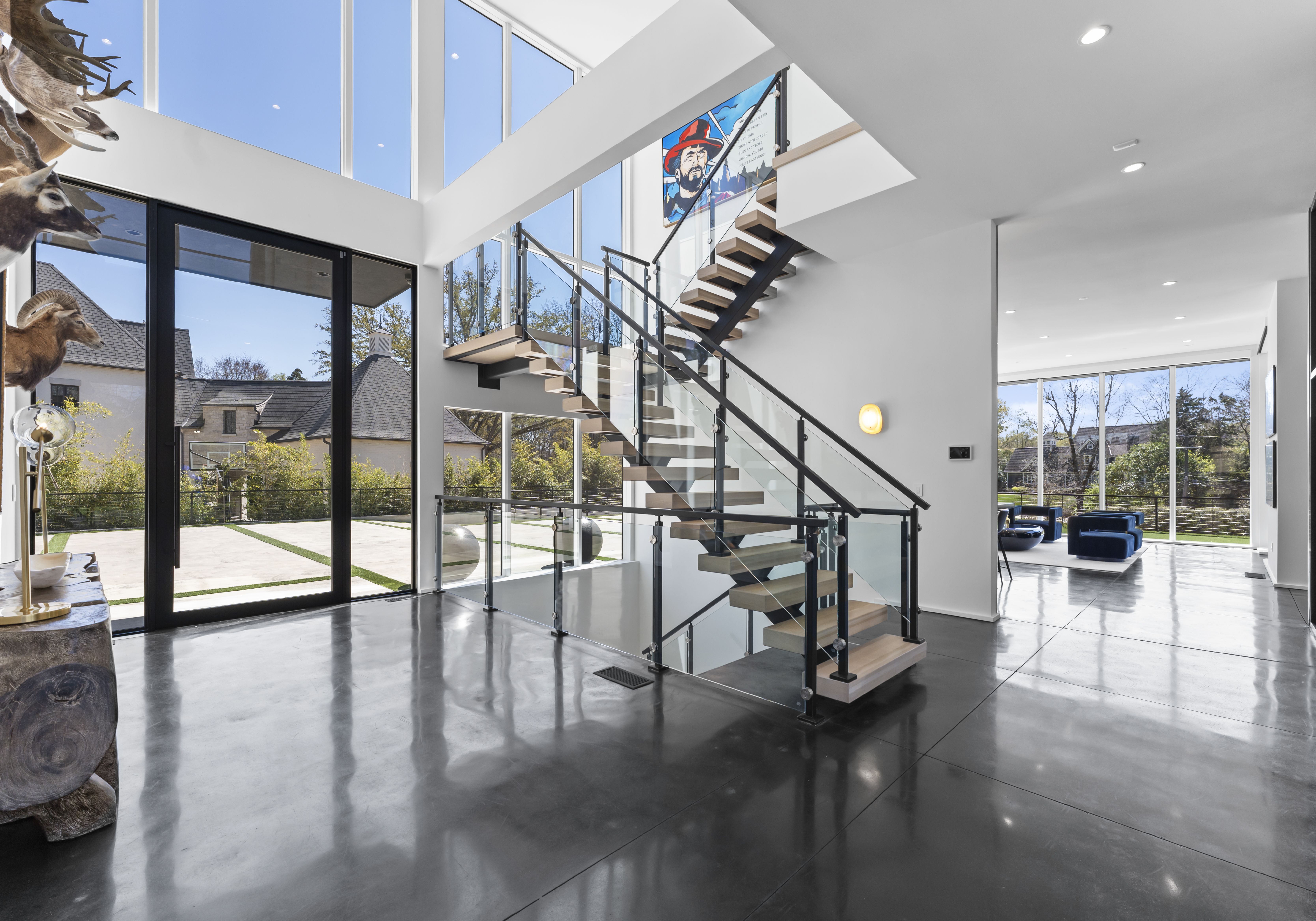 Modern home interior with floating wooden stairs and glass railings, polished concrete floor, large windows showing a sunny yard, blue chairs in living room, and animal head mounts.