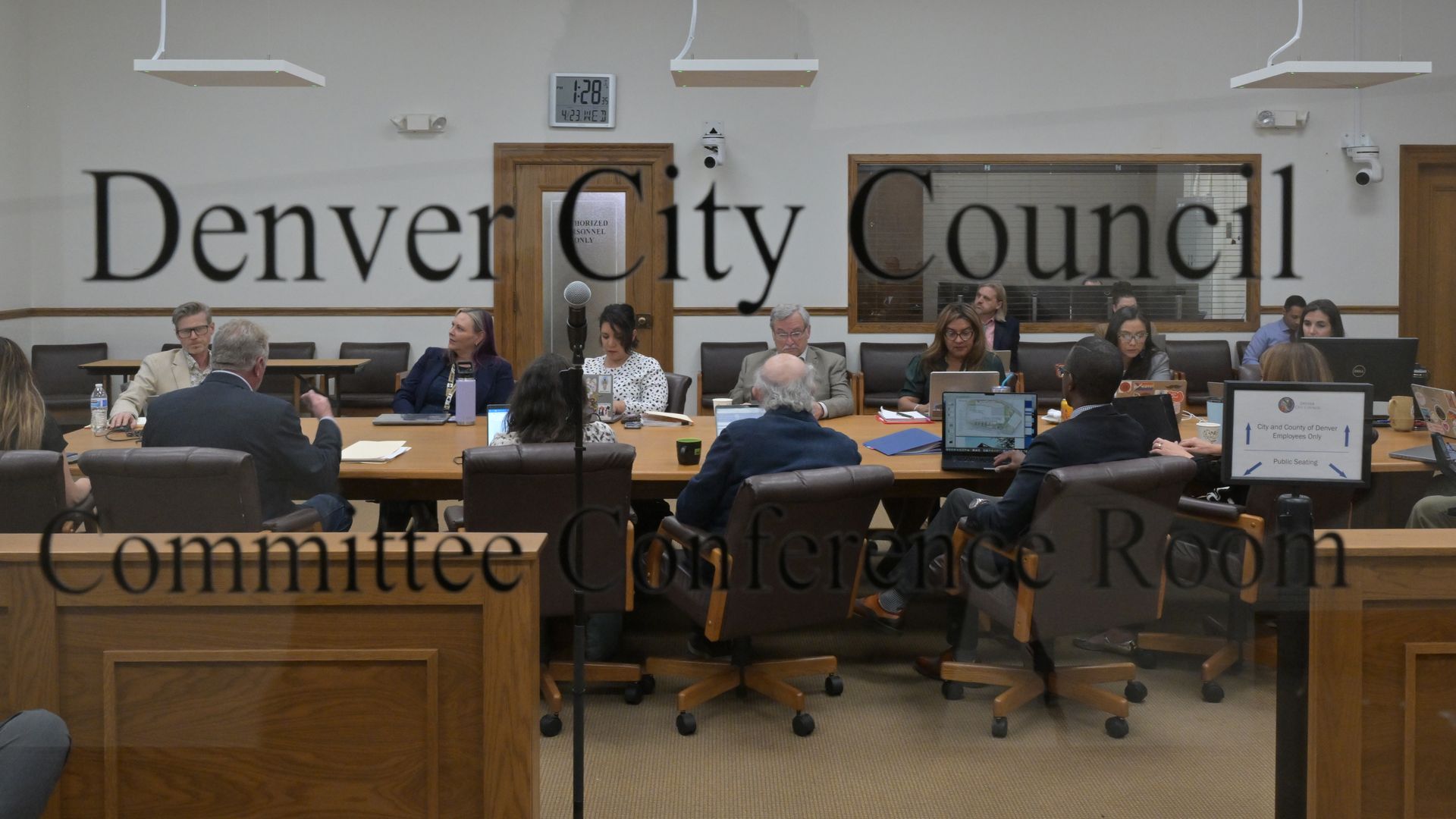 A Denver City Council meeting with officials seated around a long wooden table, laptops open, documents and beverages on the table, and a clock on the wall in the background.
