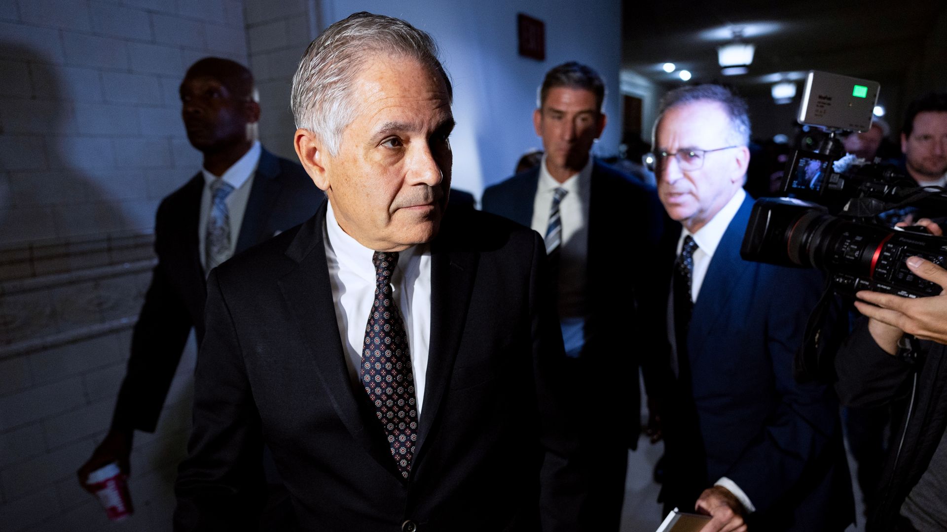 A man with white hair wearing a suit in front of several other men with cameras pointed at them walking down a hallway. 