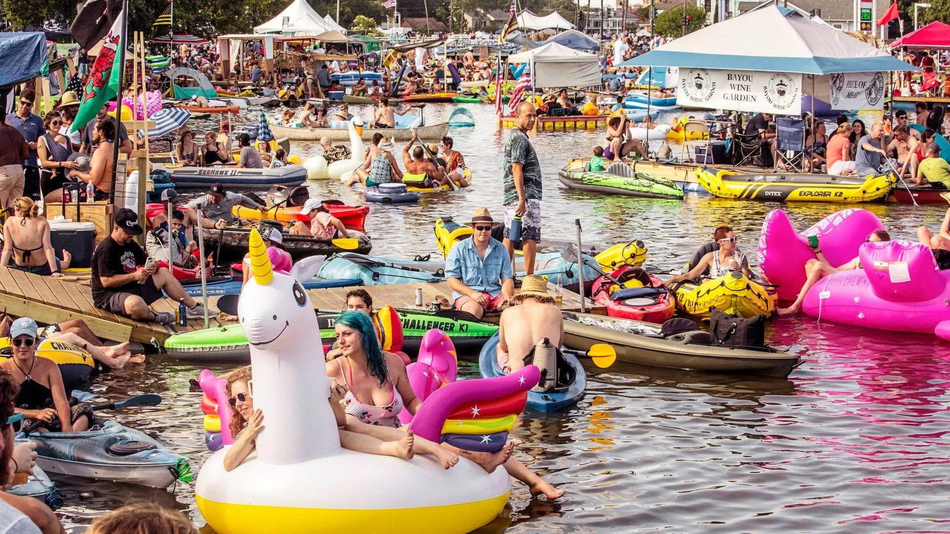 Photos shows people sitting on a giant unicorn float, swan float, flamingo float, kayaks and other rafts in Bayou St. John. Other people are on the banks.