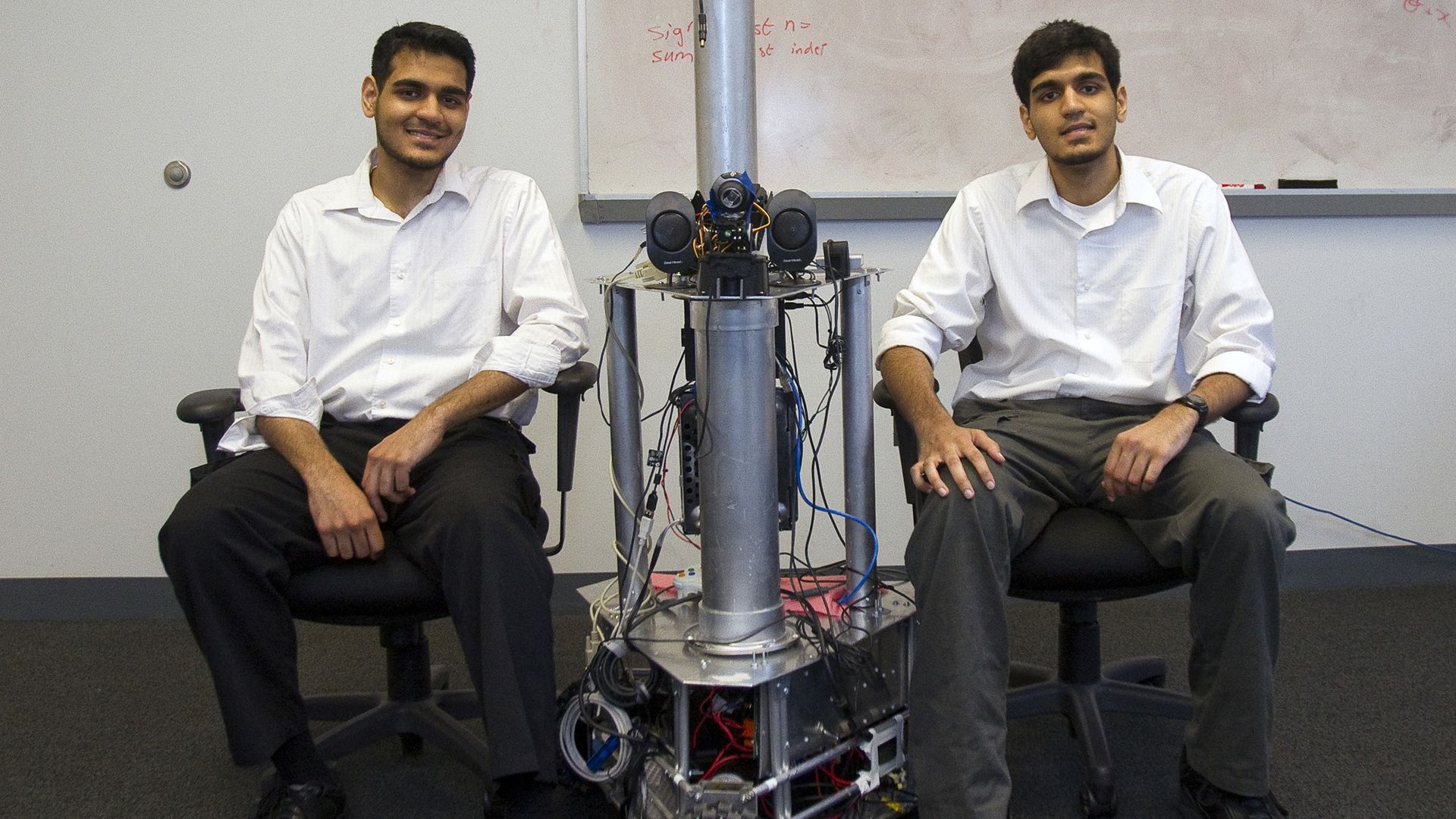 Image of Sohaib Akhter (left) and his twin brother, Muneeb, sitting down next to a geomobile omnidirectional robotic telepresence (GORT) as well as an Android based haptic vision substitution system they created  while studying at George Mason University