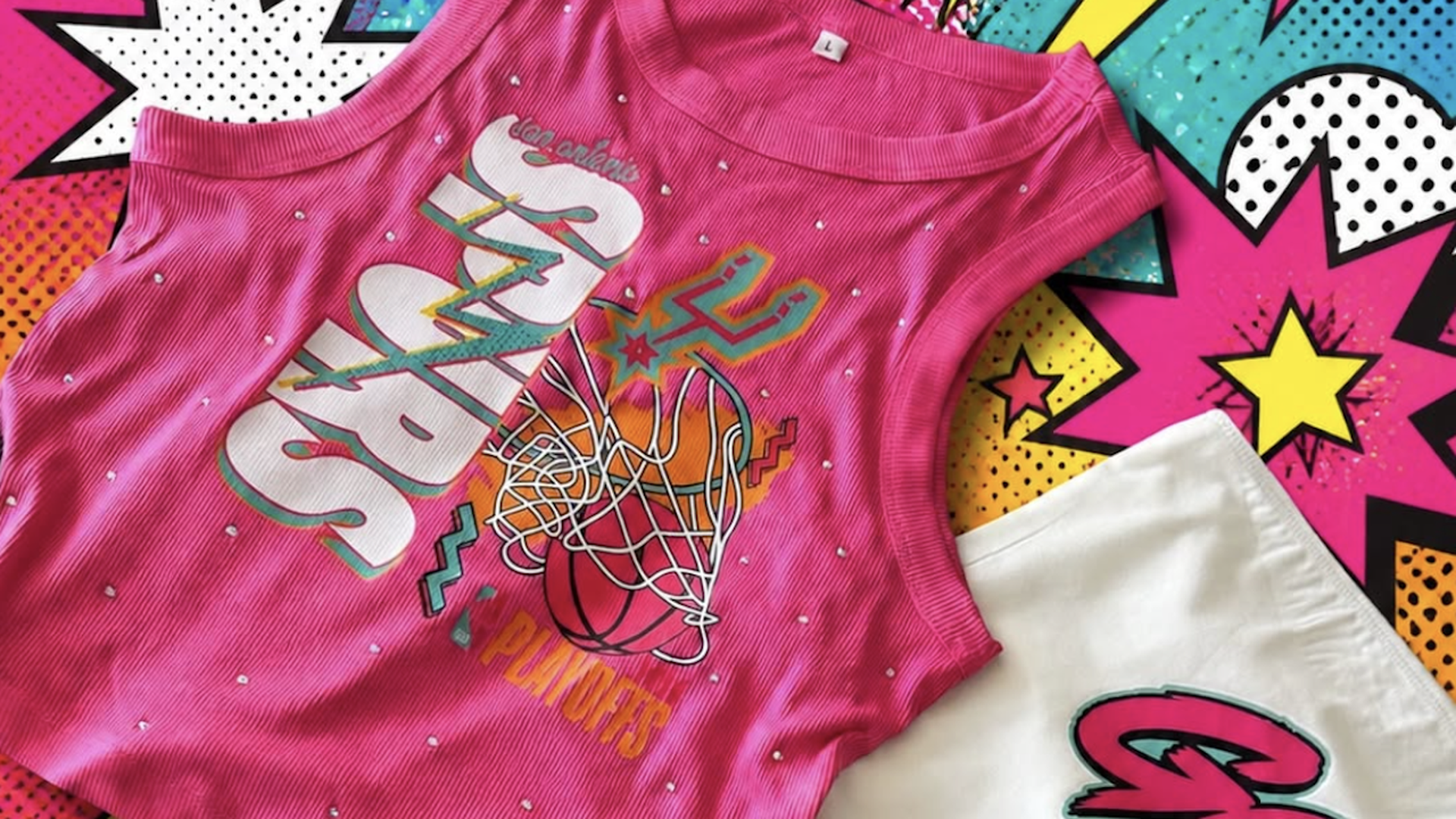 Bright pink sleeveless top with white dot pattern and a bold basketball hoop graphic, set against a vibrant pop-art background of stars and lightning bolts; a white shirt with pink lettering peeks in from the corner.