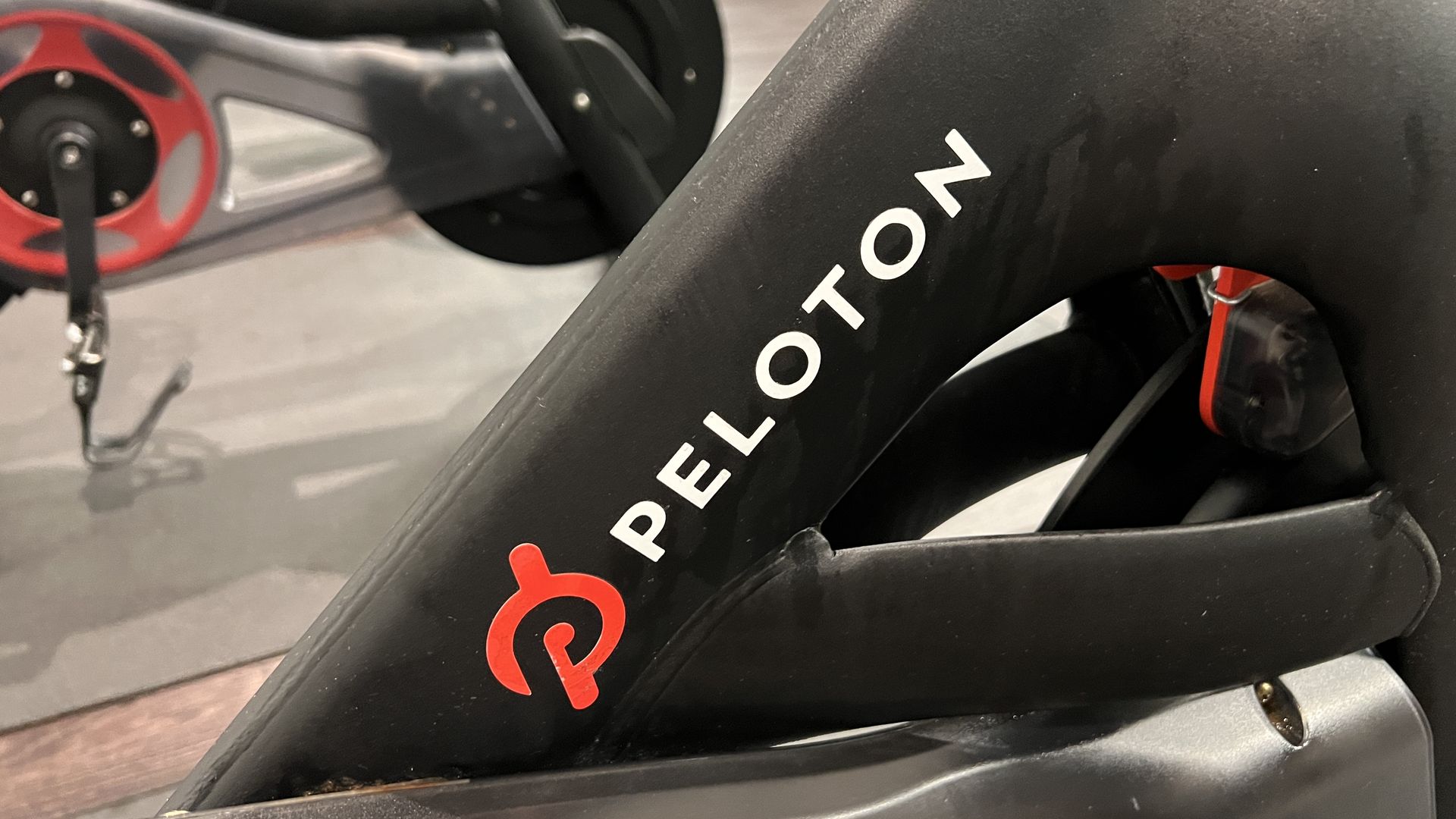 Close-up of a black Peloton stationary bike frame with white PELOTON lettering and a red logo; the frameโs triangular support is visible while a red wheel and pedal blur in the background.
