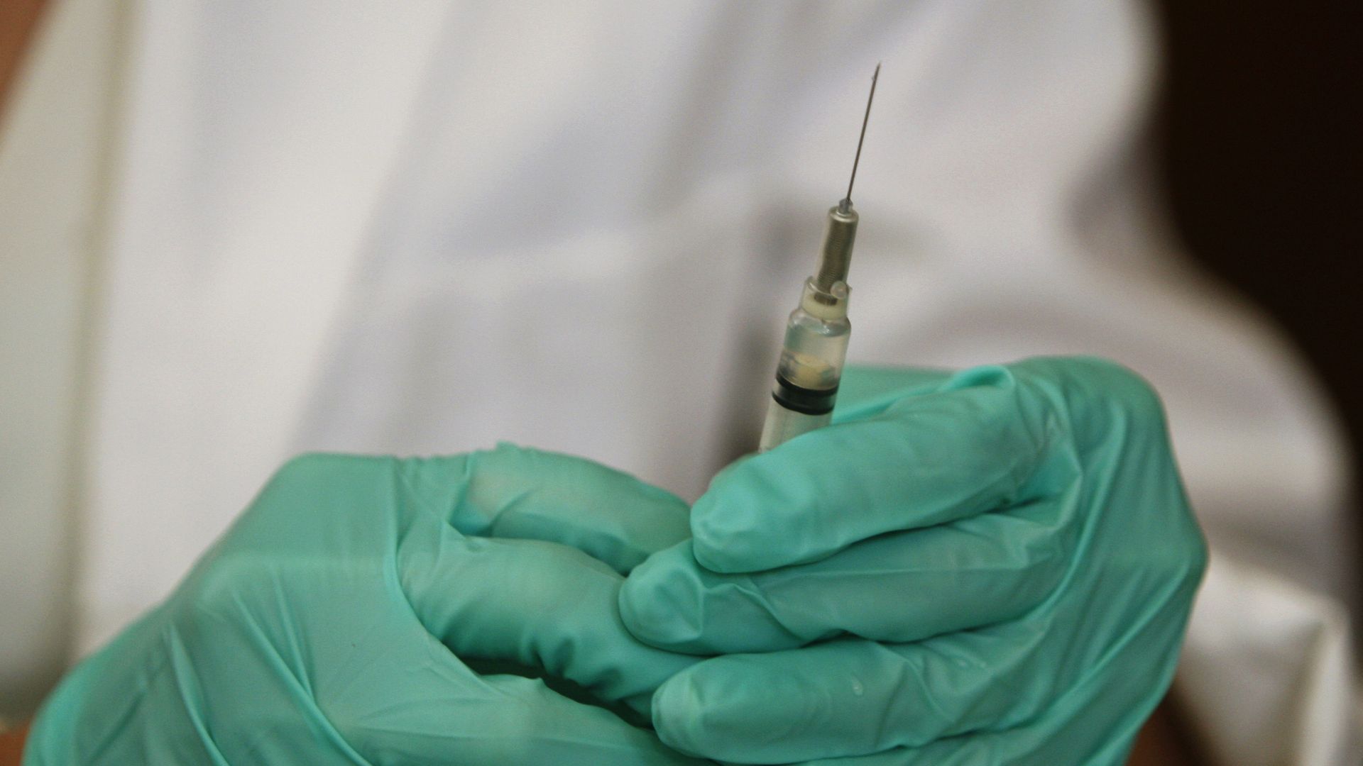 A green gloved medical provider holds a syringe of liquid. 