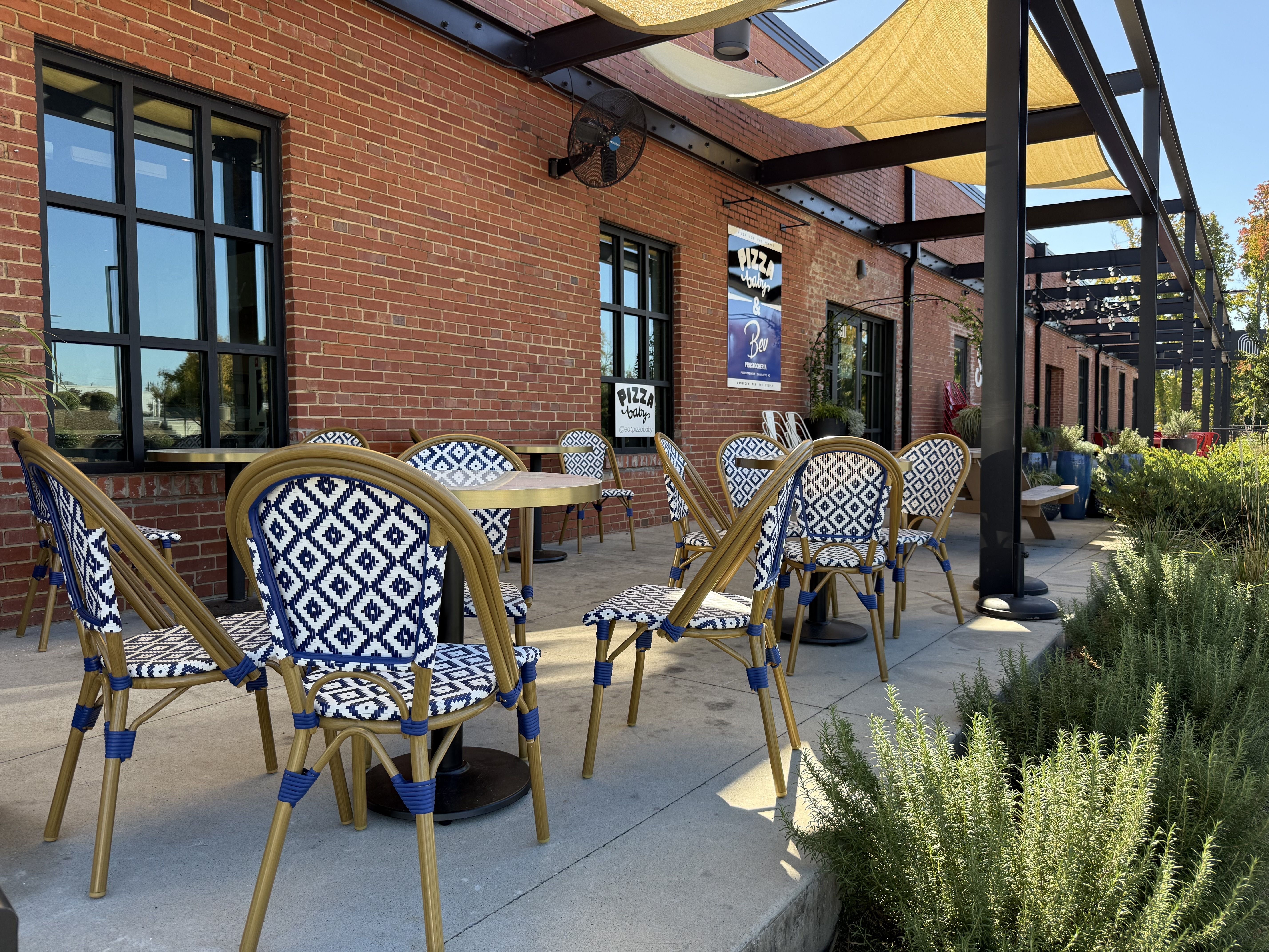 Outdoor cafe patio beside a brick building with large black-framed windows; many blue and white geometric patterned chairs around round tables under tan fabric shade sails.