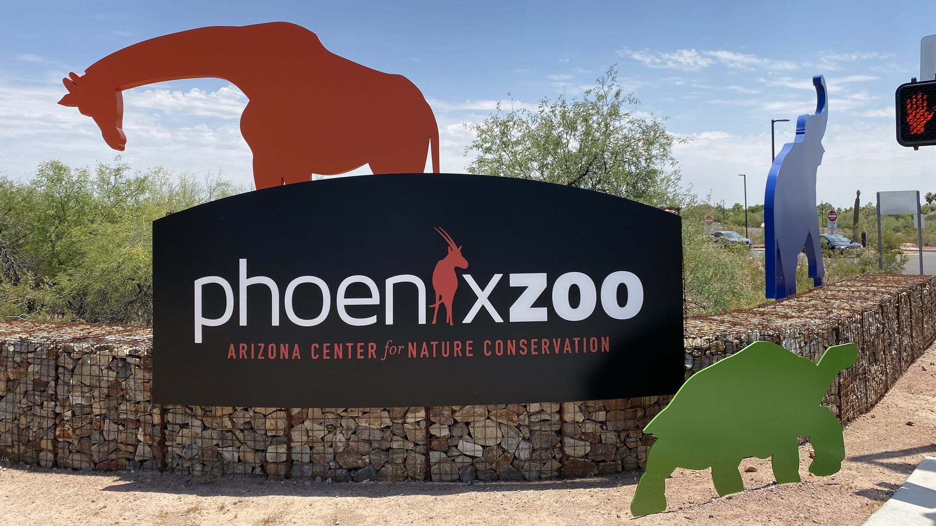 Flat metal red, green and blue sculptures shaped like a giraffe turtle and elephant next to a sign that reads Phoenix Zoo.