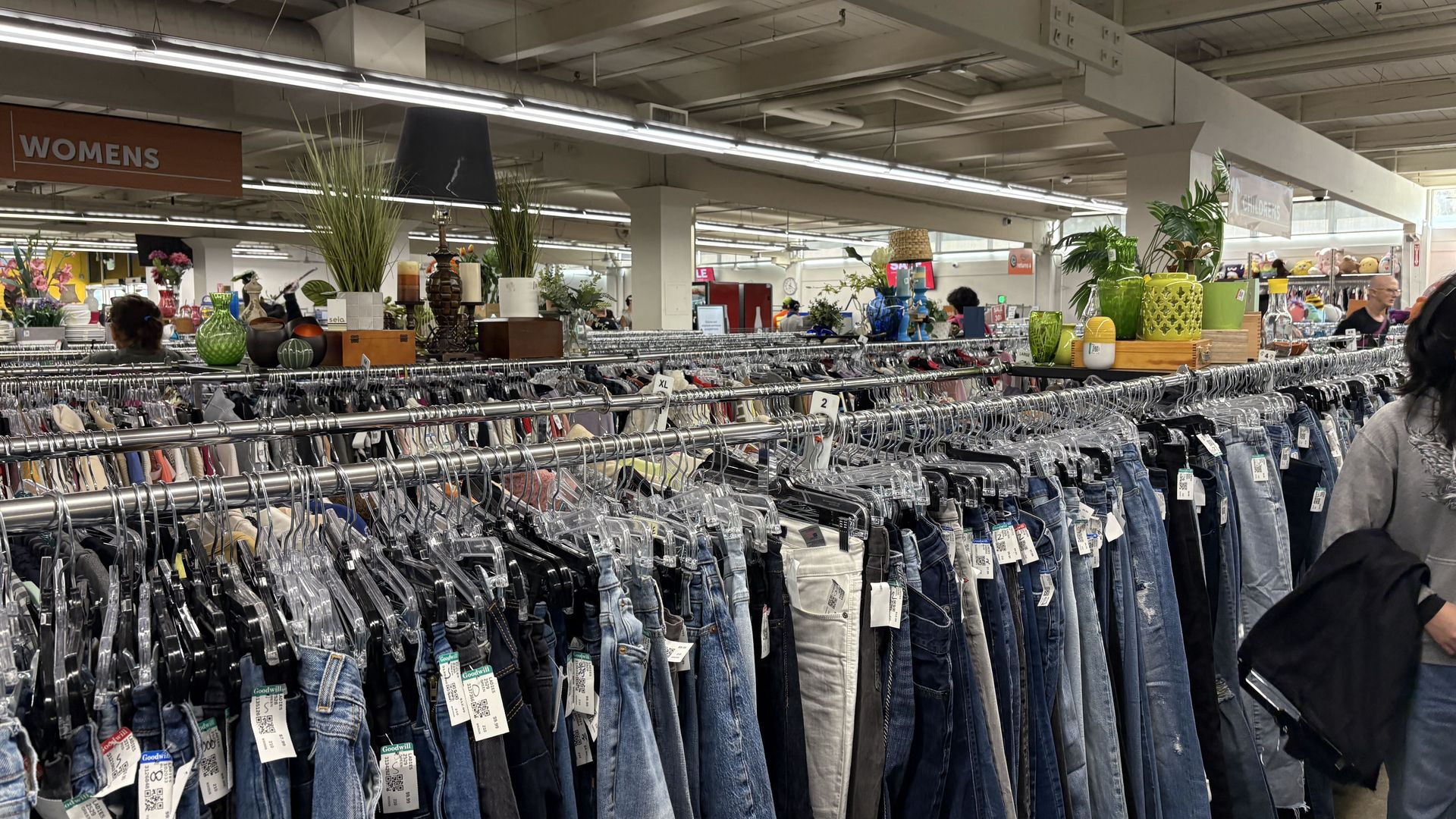 Interior of a large thrift store with denim jeans on metal racks in the foreground, a "WOMENS" sign to the left, exposed ceiling beams and bright fluorescent lights, and shoppers in the background.