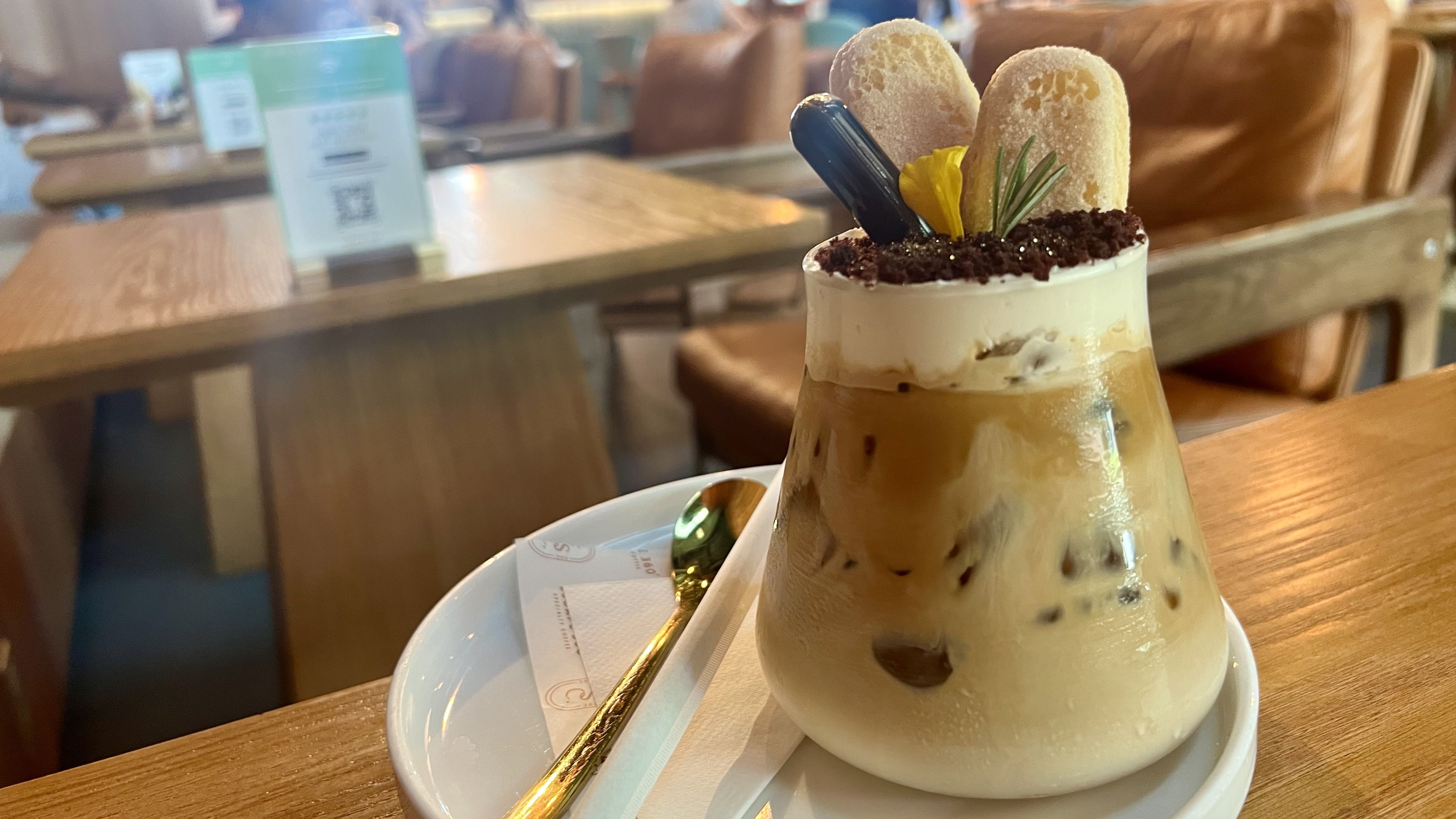 Iced coffee drink in a glass with whipped cream, two ladyfinger biscuits, a yellow flower, and herbs on top, served on a white plate with a gold spoon on a wooden table in a cozy cafe.