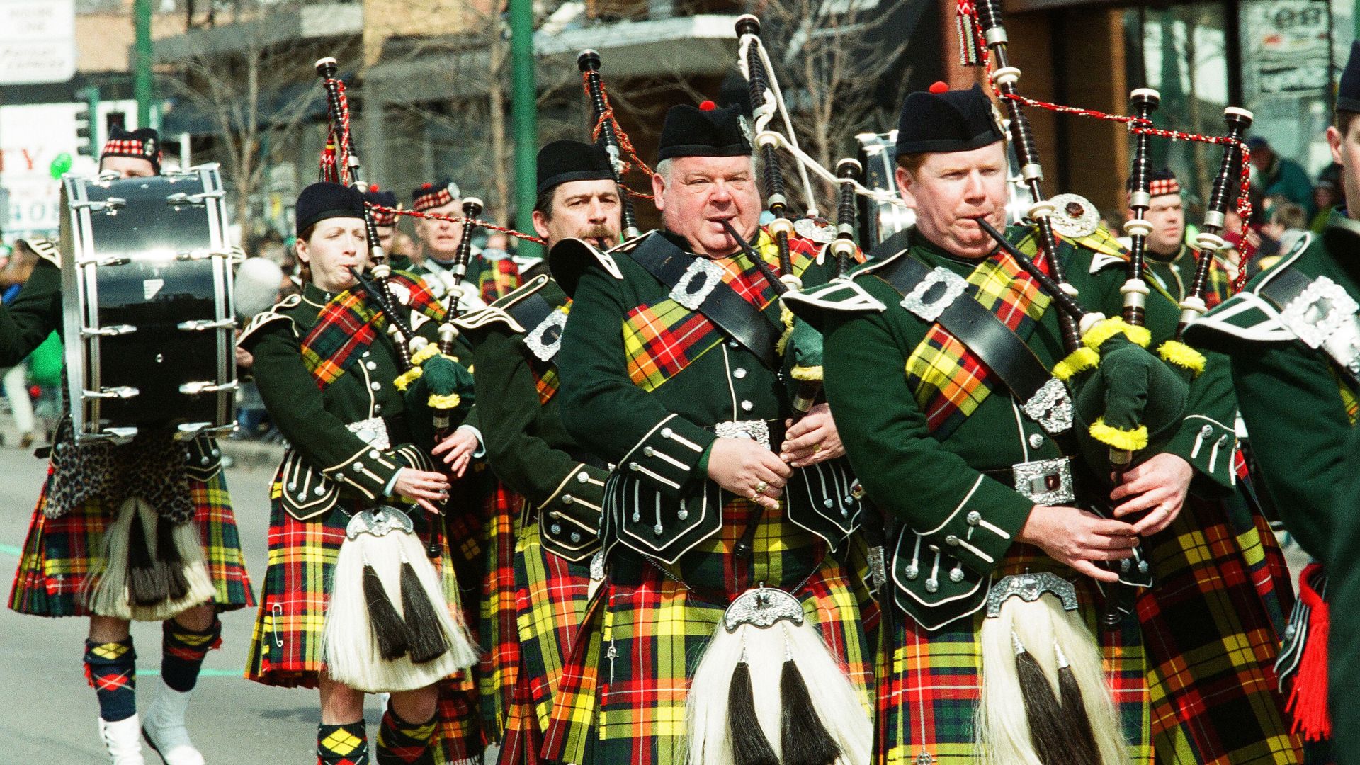 Parade of bagpipers in green jackets and bright plaid kilts, playing bagpipes and drums as they march down a city street with spectators and storefronts in the background.