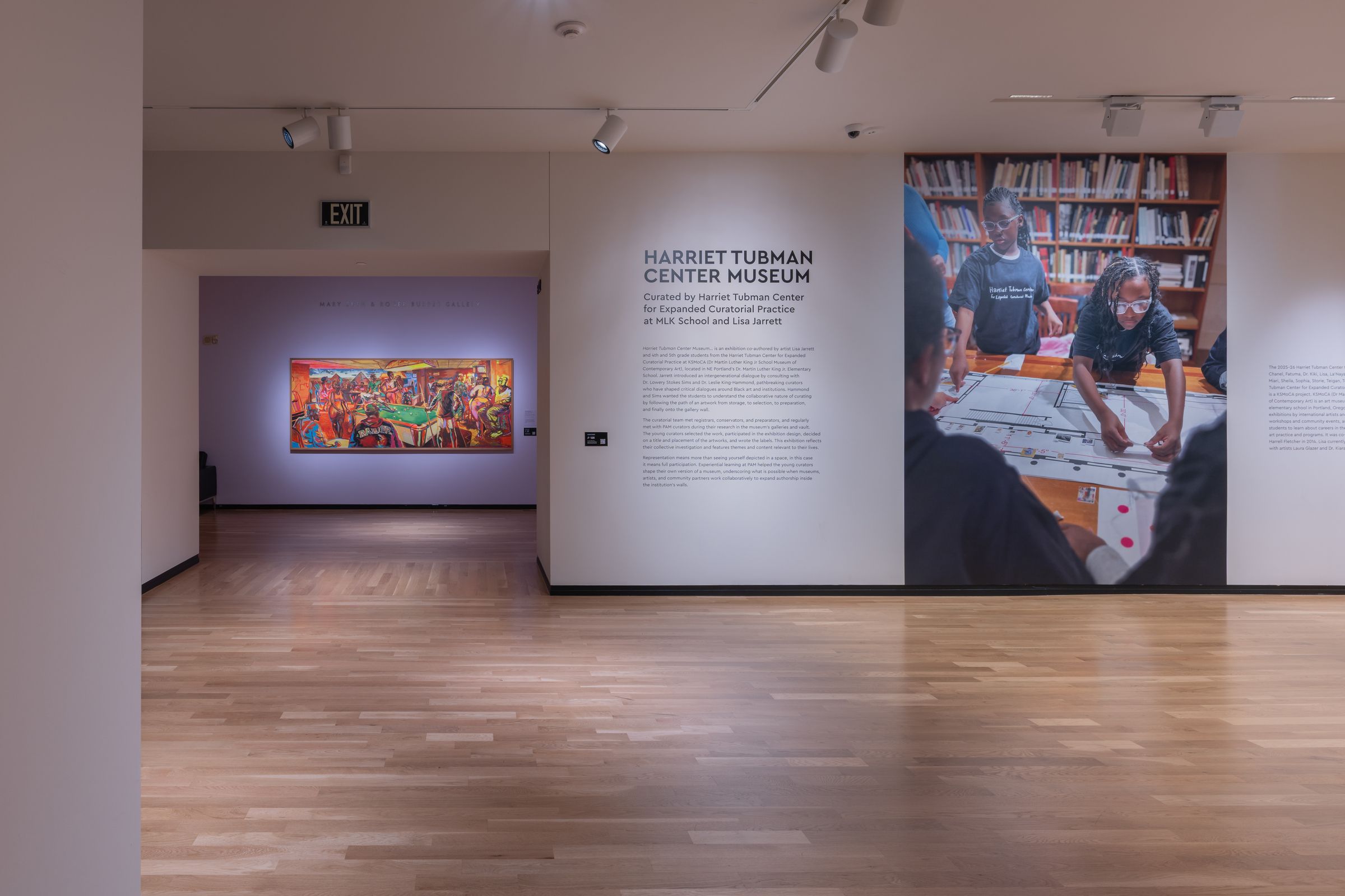 Museum hallway with wood floor; central wall reads "HARRIET TUBMAN CENTER MUSEUM." Right shows a large library photo; left a purple gallery with a colorful painting; exit sign overhead.