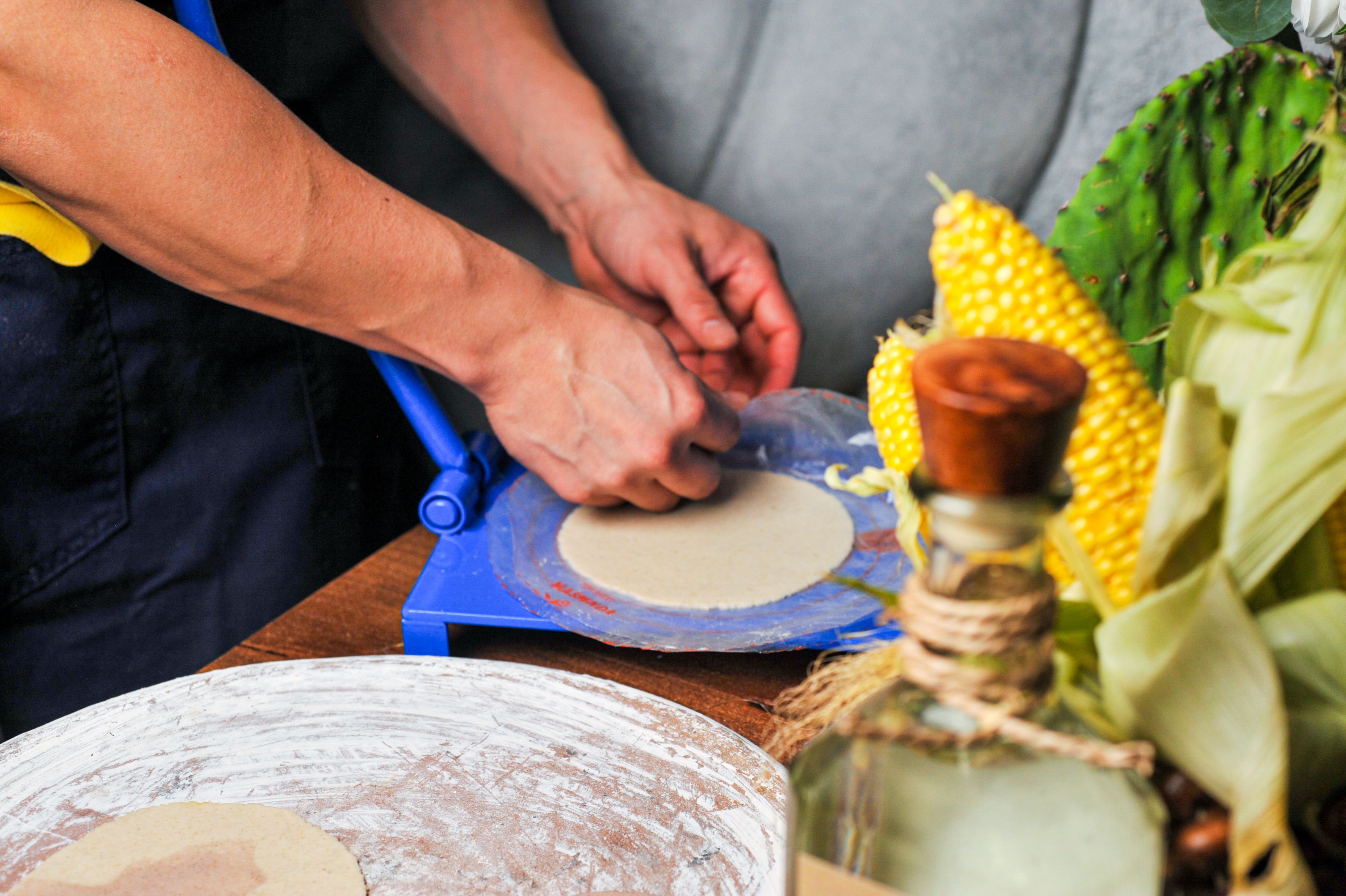 Person's hands operate a blue tortilla press, shaping masa disks; in the foreground are corn on the cob and green husks on a wooden surface.