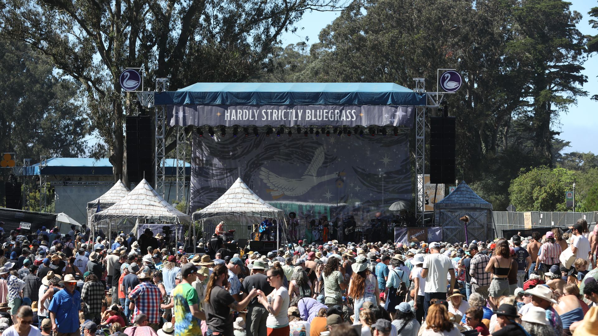 Outdoor festival stage with a blue canopy and a banner reading "Hardly Strictly Bluegrass." A packed crowd wearing hats fills the foreground, with tents in front and tall trees behind.