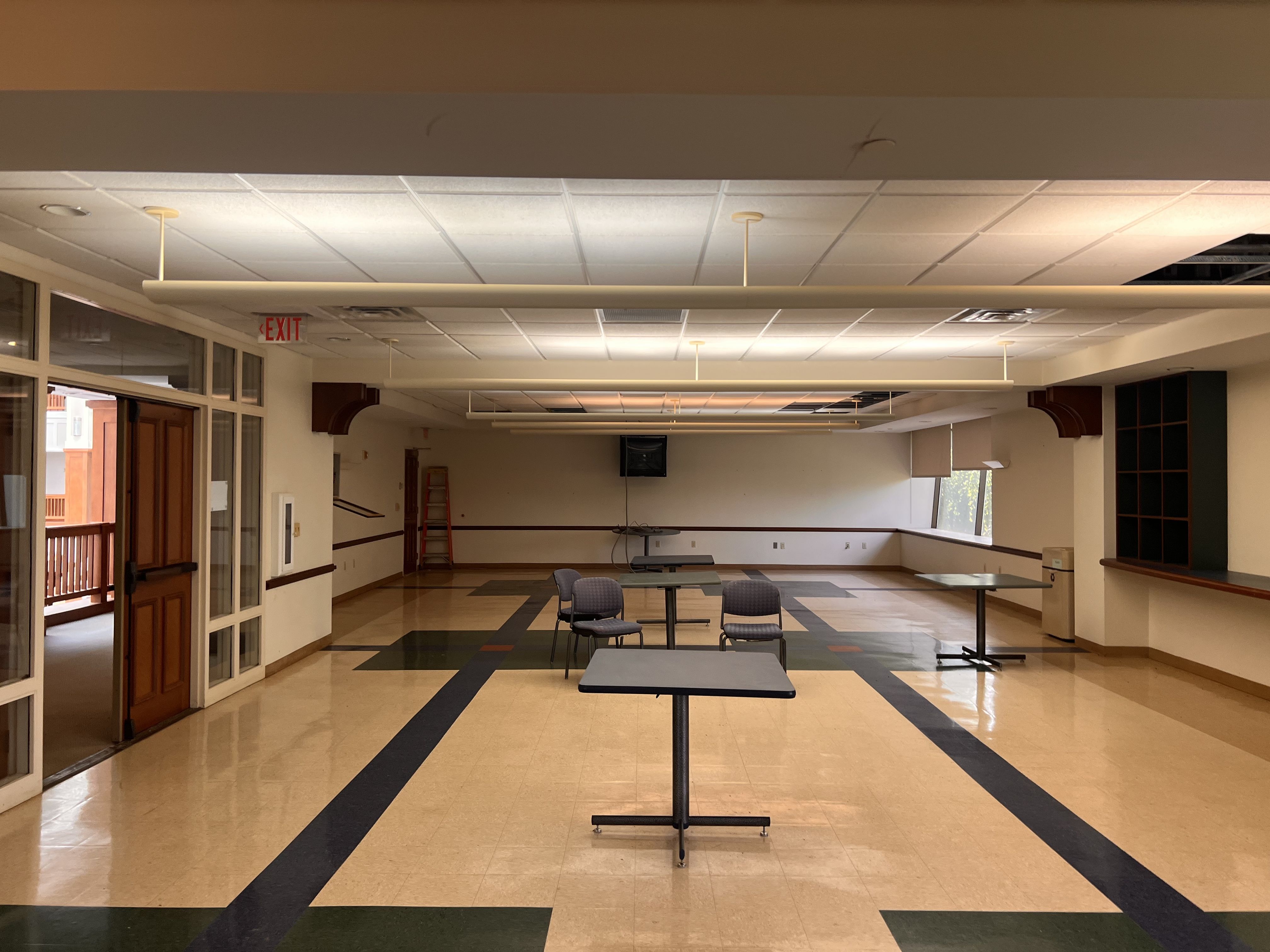 Interior hall with tiled floor, long white pendant lights along a dropped ceiling. Glass doors and an EXIT sign on the left; scattered tables and chairs, ladder in back.