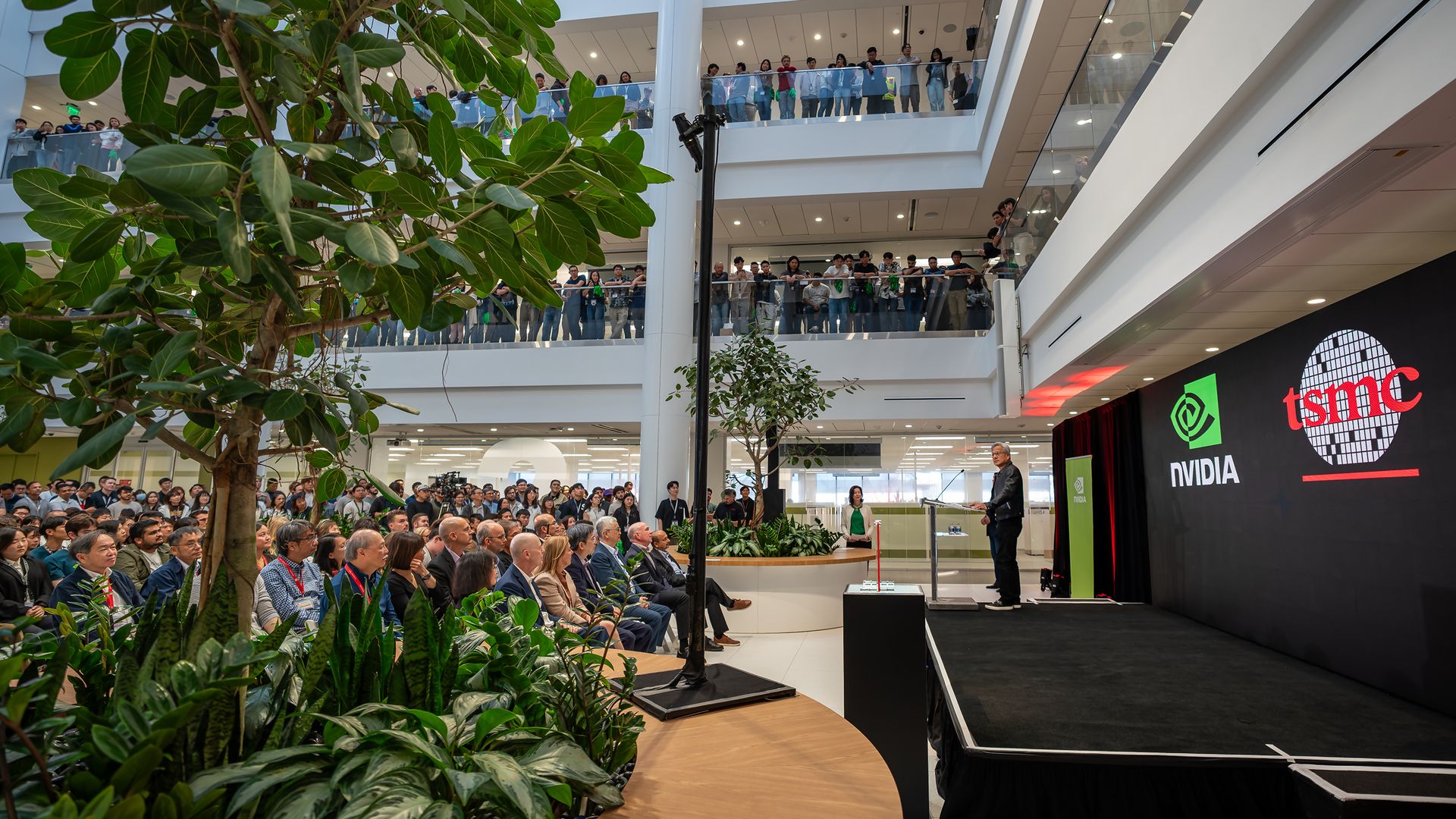 A large crowd watches a speaker on stage in a multi-level modern atrium. Logos for NVIDIA and TSMC are displayed on a screen behind the speaker. Green plants decorate the space.