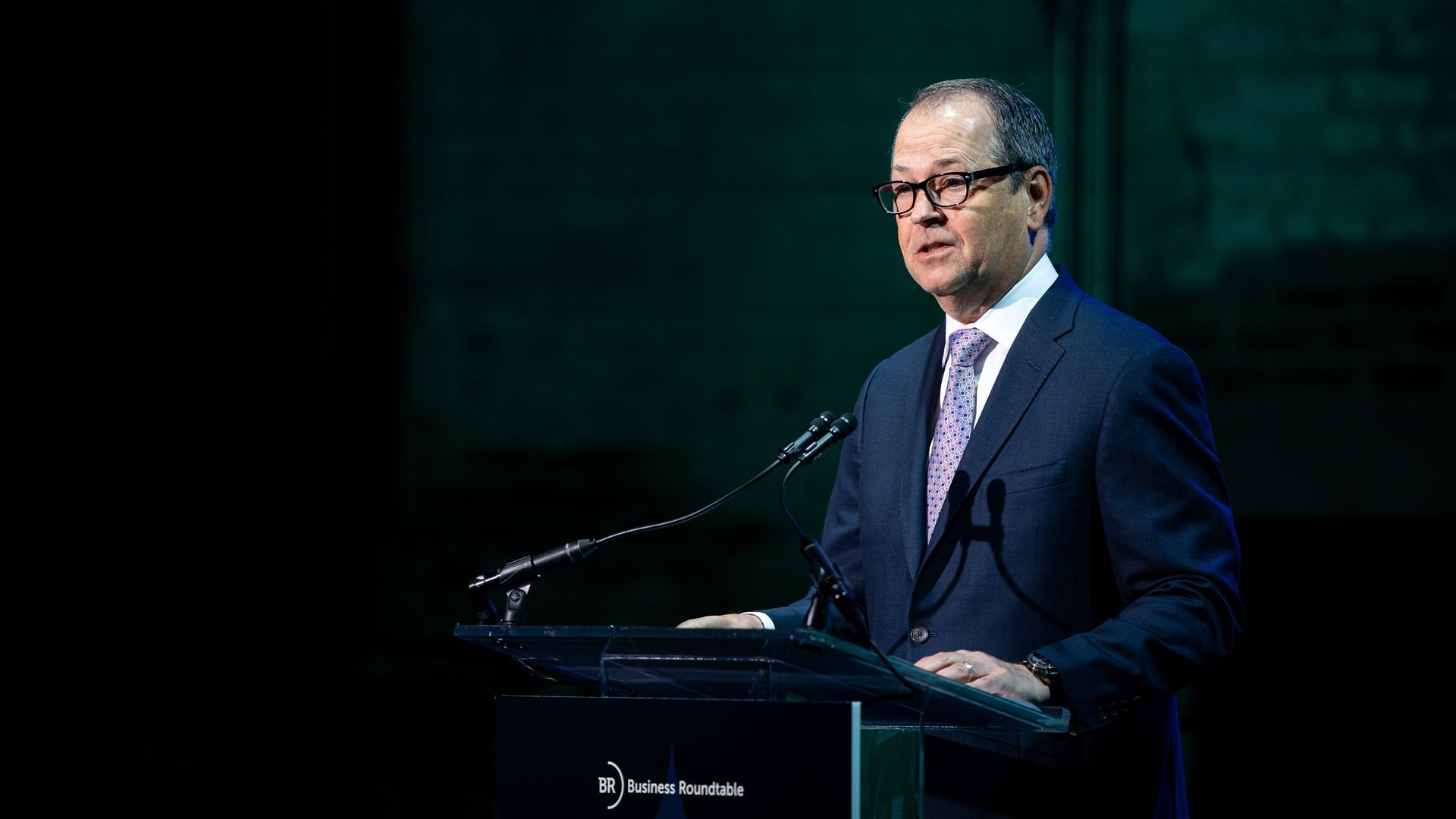 Man in navy suit and patterned tie speaking at a clear podium with Business Roundtable logo, against a dark green blurred background.
