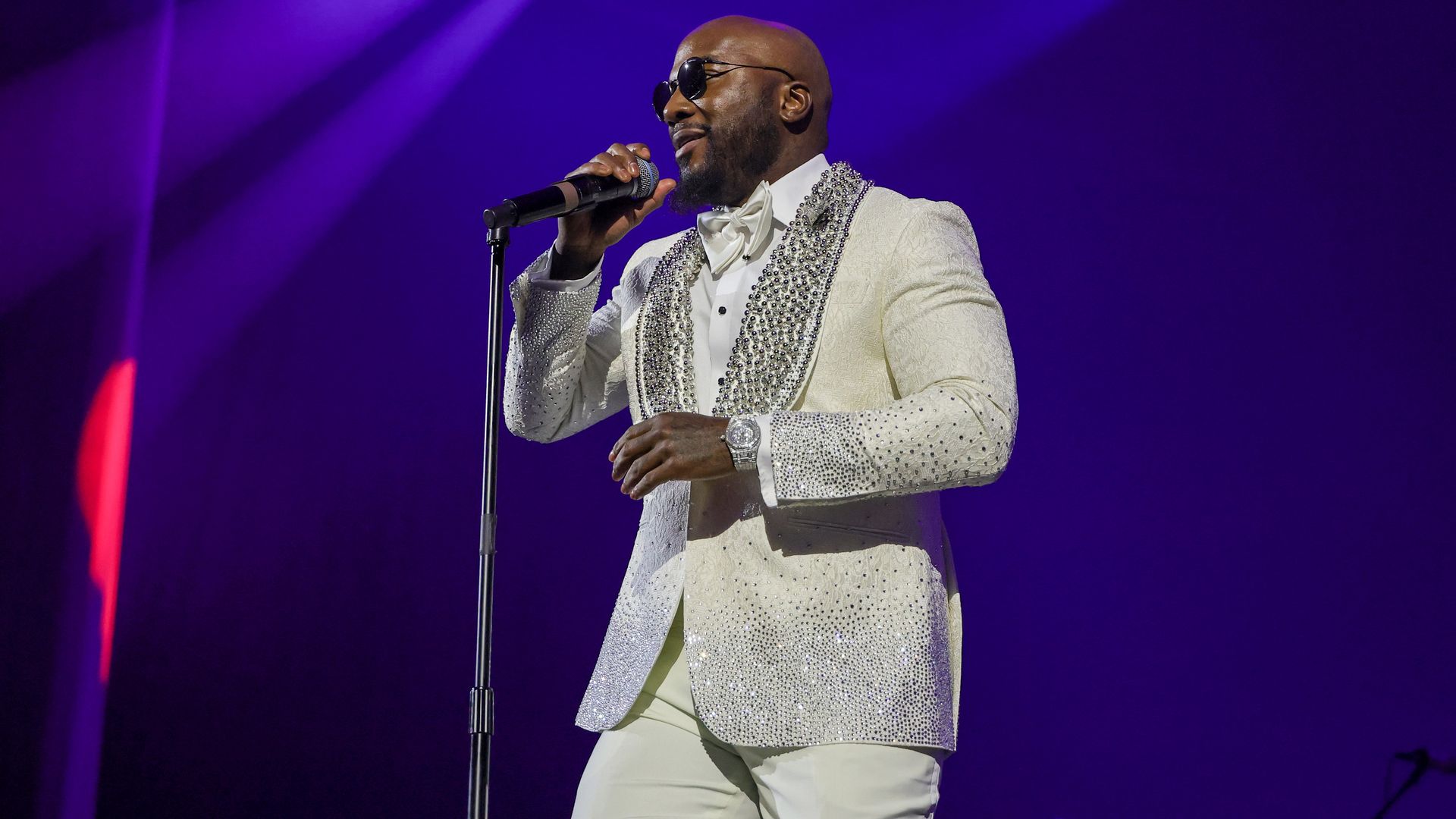 A man wearing sunglasses, diamond watch and a cream-colored suit adorned with diamonds holds a microphone in his right hand as he sings on stage. 