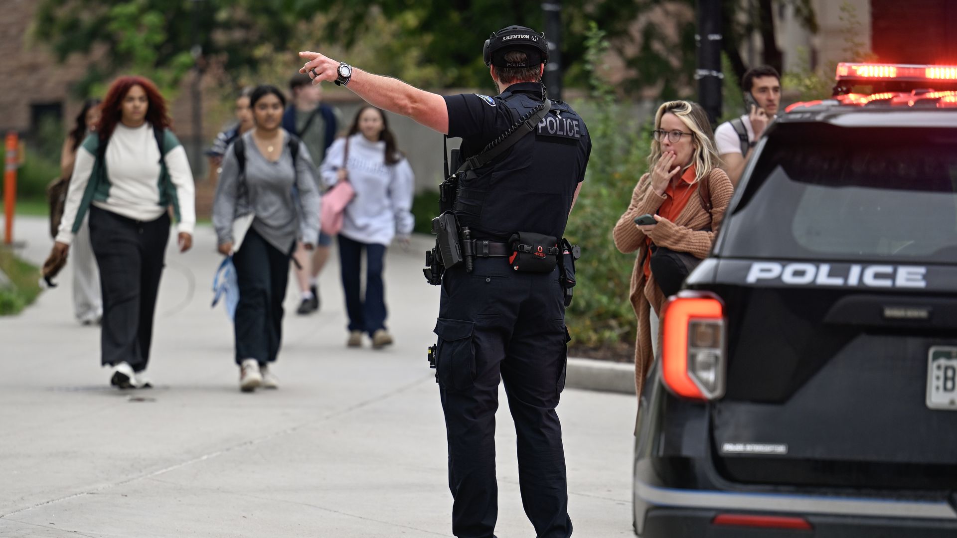 Police officer in black uniform directs or stops group of people walking on sidewalk beside police vehicle with flashing lights, while woman in brown sweater looks on holding phone.