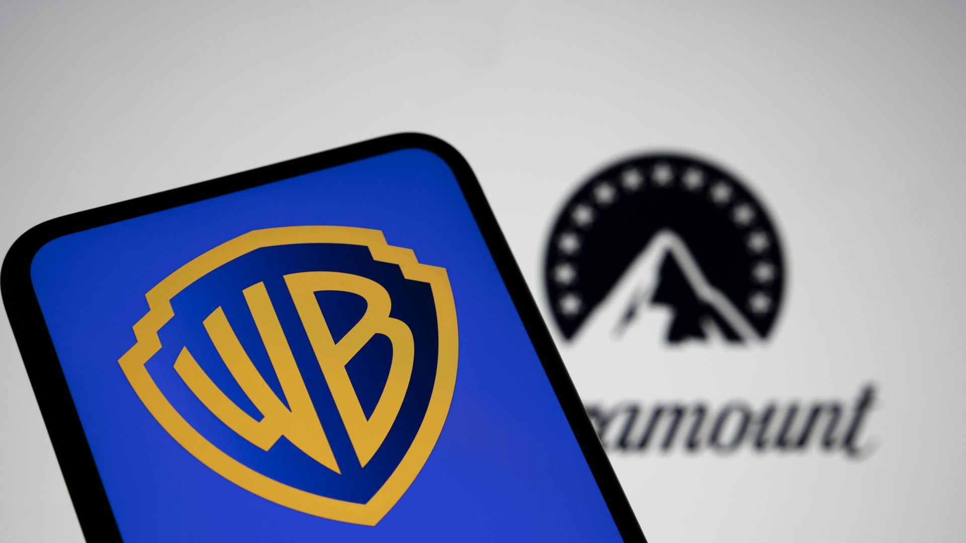 Close-up of a blue device screen displaying the yellow Warner Bros shield logo; in the blurry background is a black circular emblem with a white mountain, and faint text.