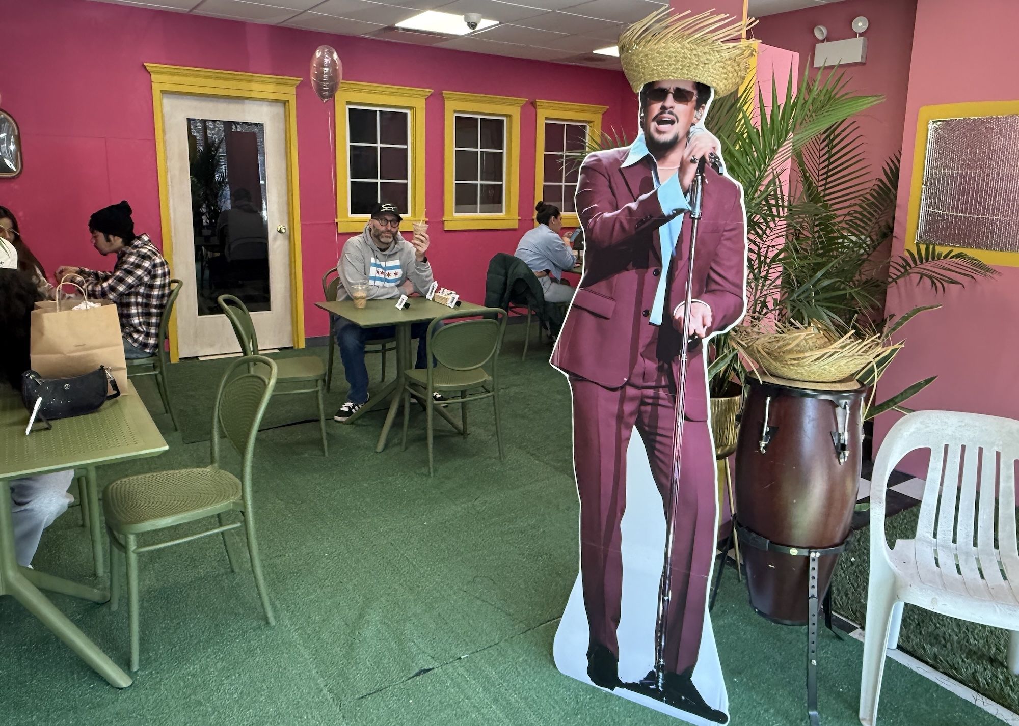 Indoor cafe with green tables and chairs, pink walls with yellow window frames, a man sitting and holding an ice cream, and a cardboard cutout of Bad Bunny in a maroon suit and straw hat.
