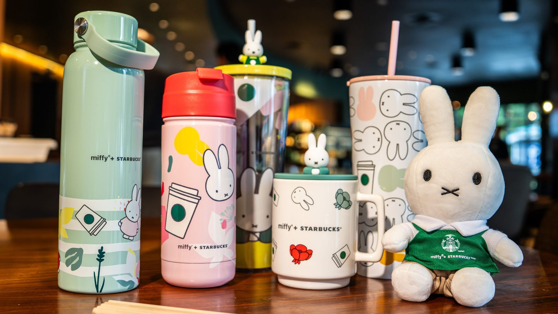 Assorted Miffy-themed Starbucks cups and a plush Miffy toy on a cafe counter. Left a mint water bottle, center pastel pink cup, right white cup with bunny patterns; pencils in front.