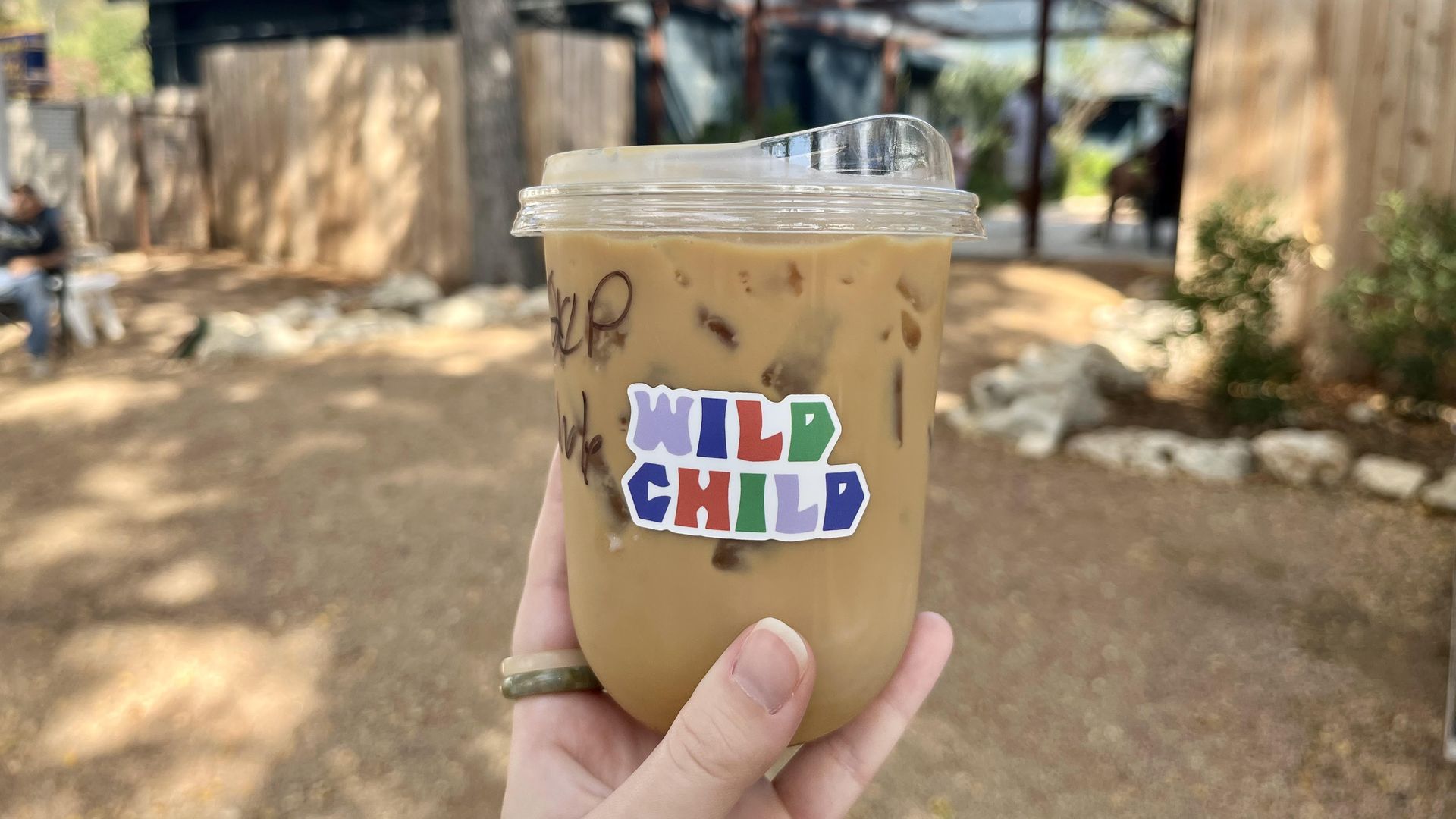 A hand holding an iced latte in a clear plastic cup with a colorful "WILD CHILD" sticker. In the background is a blurred version of the Ranch Motel outdoors with trees, wooden fences and buildings.
