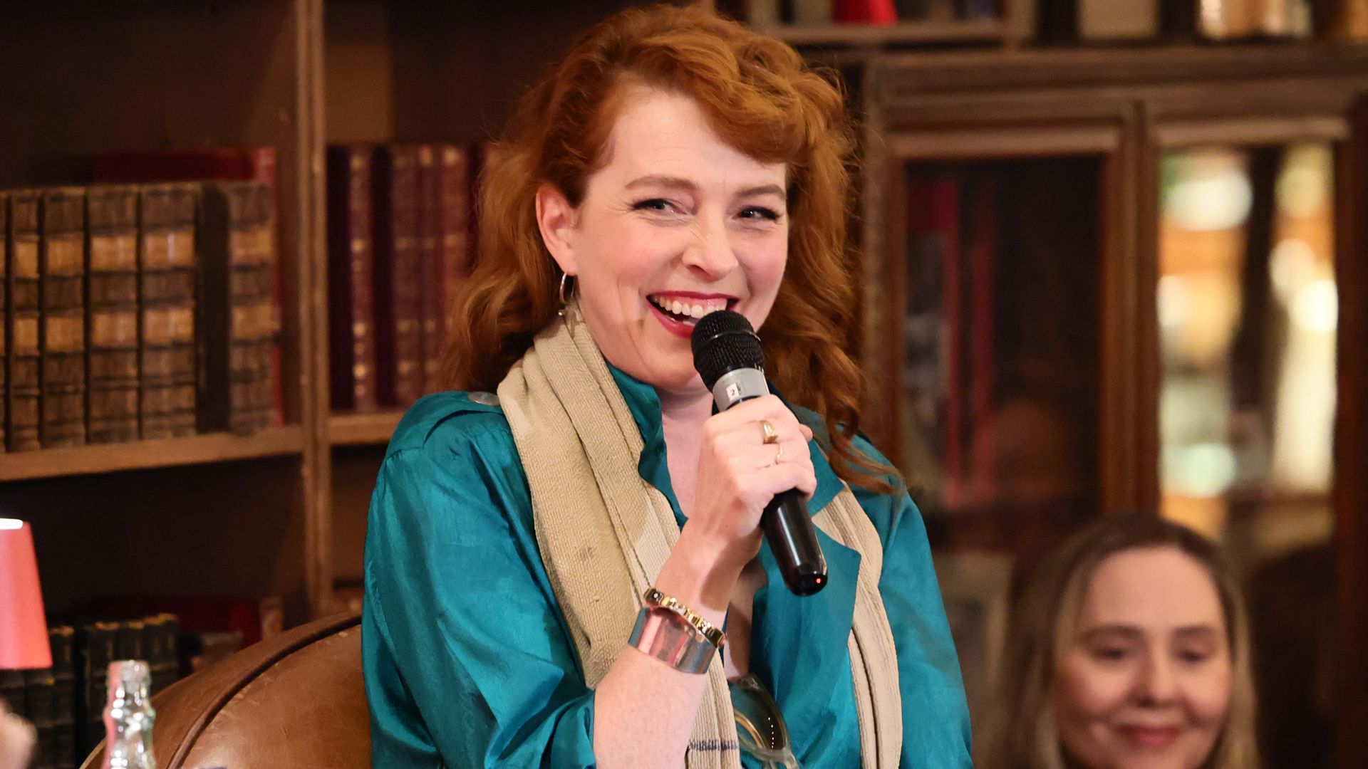 Smiling red-haired woman in a teal blouse and beige scarf holds a microphone, with glasses hanging from her scarf, in a library with wooden bookshelves behind her.