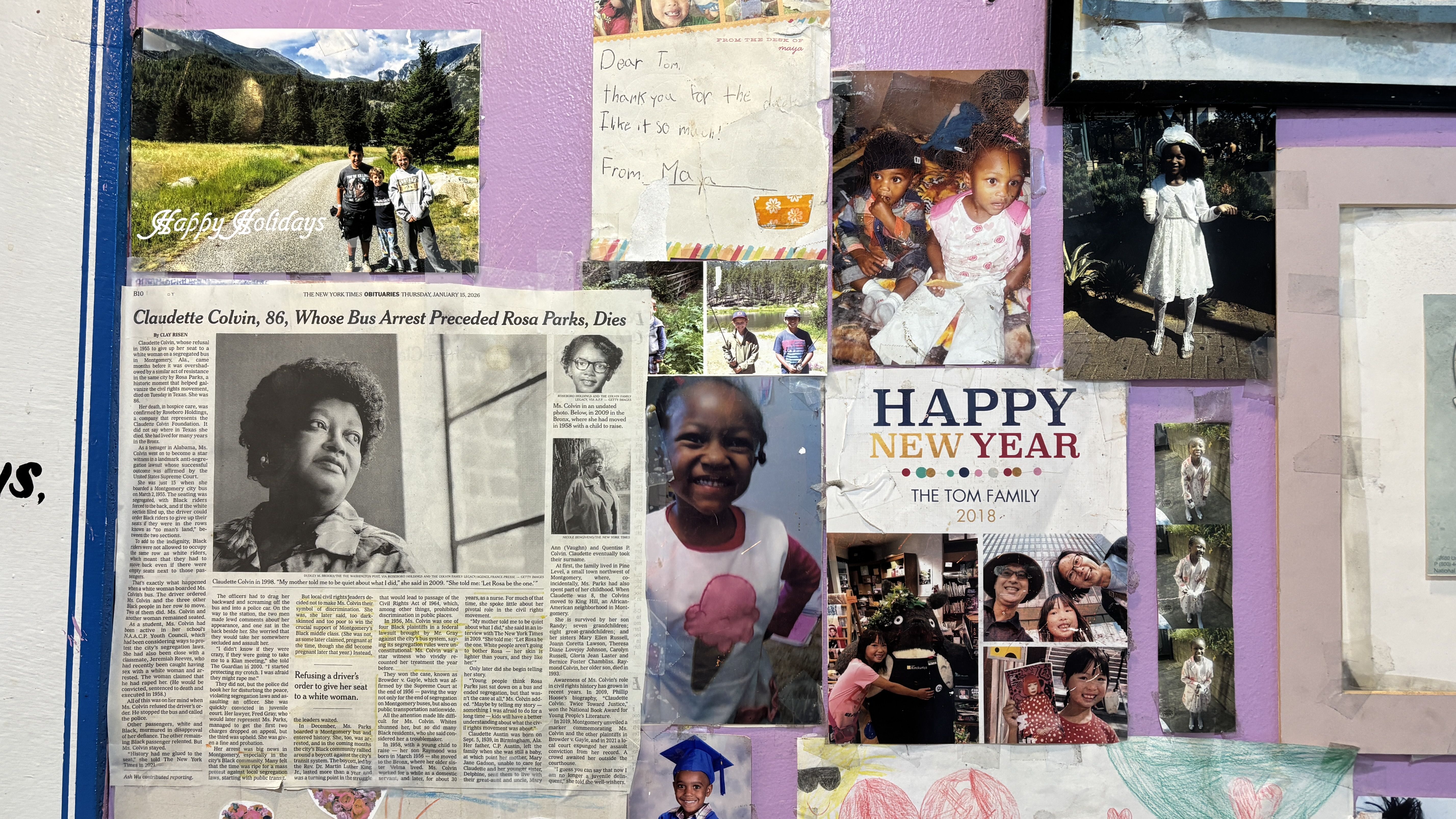 A purple wall covered with various family photos, children's drawings, a newspaper obituary for Claudette Colvin, a Happy Holidays card, and a Happy New Year 2018 message from the Tom family.