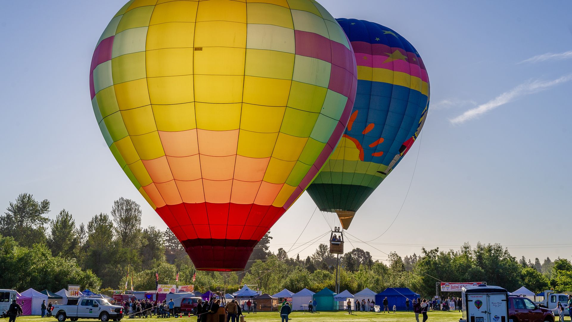 Two large, colorful hot air balloons float over a grassy festival field with tents, parked vehicles, and people watching; clear blue sky and trees in the distance.
