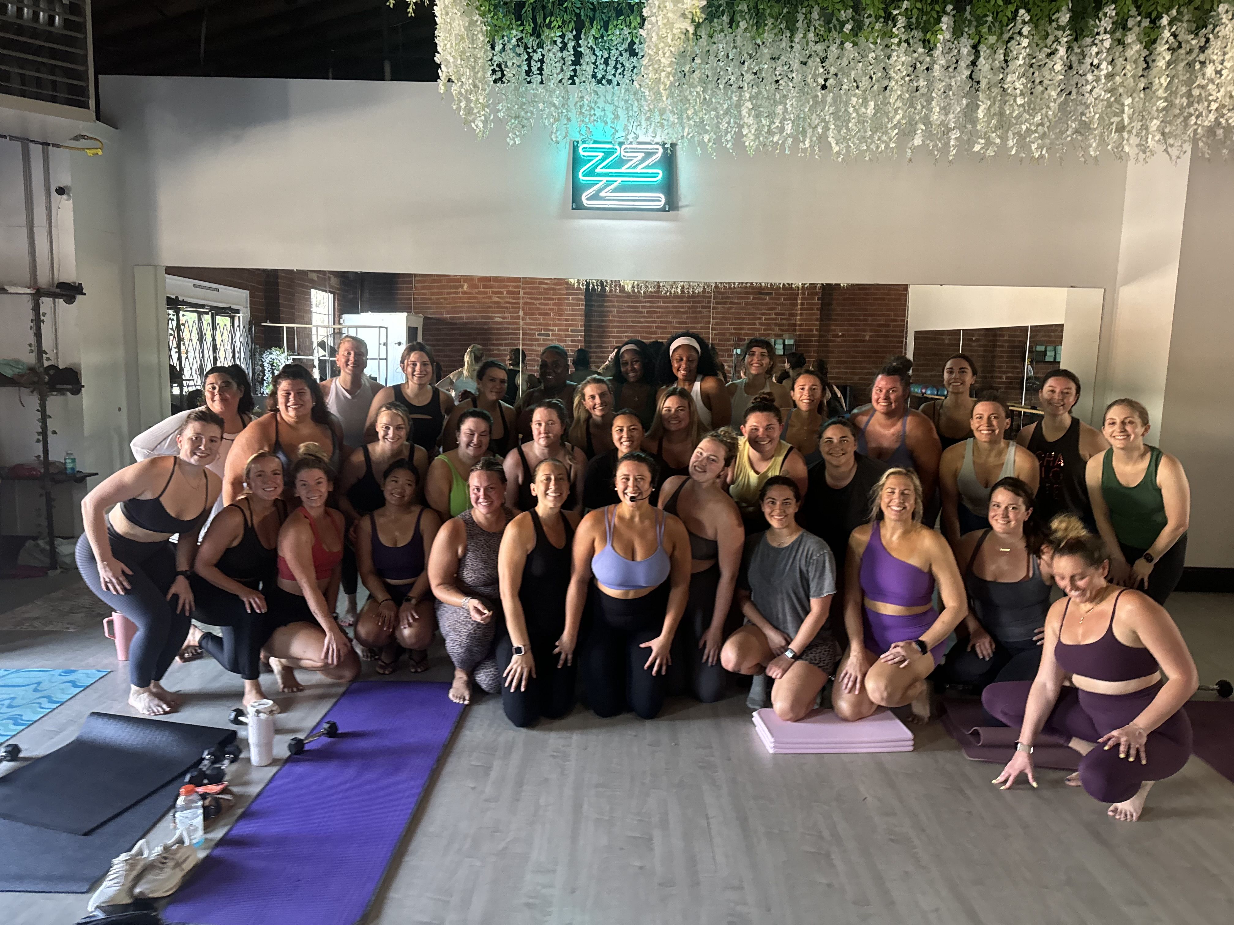 Group of diverse women in athletic wear posing and smiling in a fitness studio with yoga mats, weights, and a faux floral ceiling, under a neon ZZZ sign.
