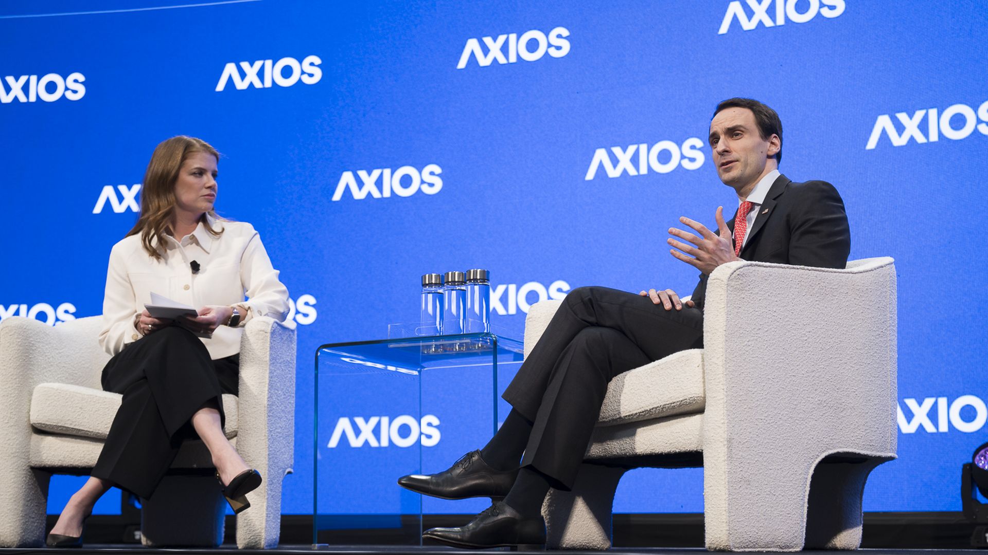 Two people sit on a stage in white armchairs with a blue Axios backdrop. A person in a white blouse and black pants holds papers; a person in a dark suit and red tie gestures as he speaks.