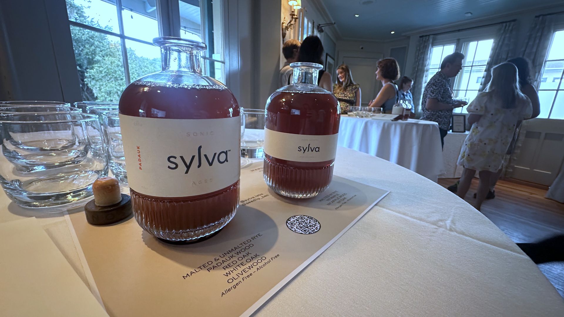 Two brown glass bottles labeled "sylva" on a white tablecloth with empty glass cups nearby; people chat in a softly lit room with large windows in the background.