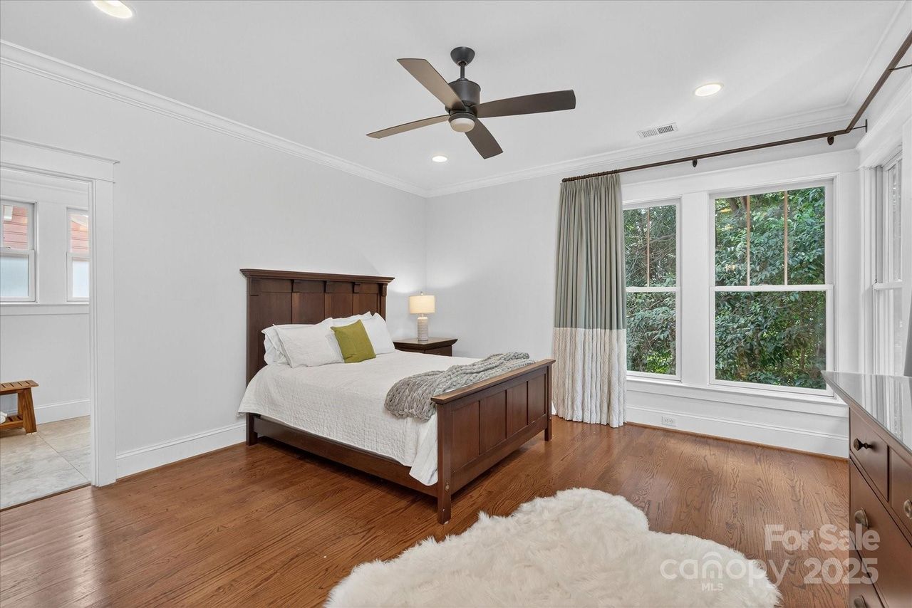 Bedroom with wooden bed frame and white bedding, green pillow, nightstand with lamp, wood floor, large window with green and white curtains, ceiling fan, and white walls.