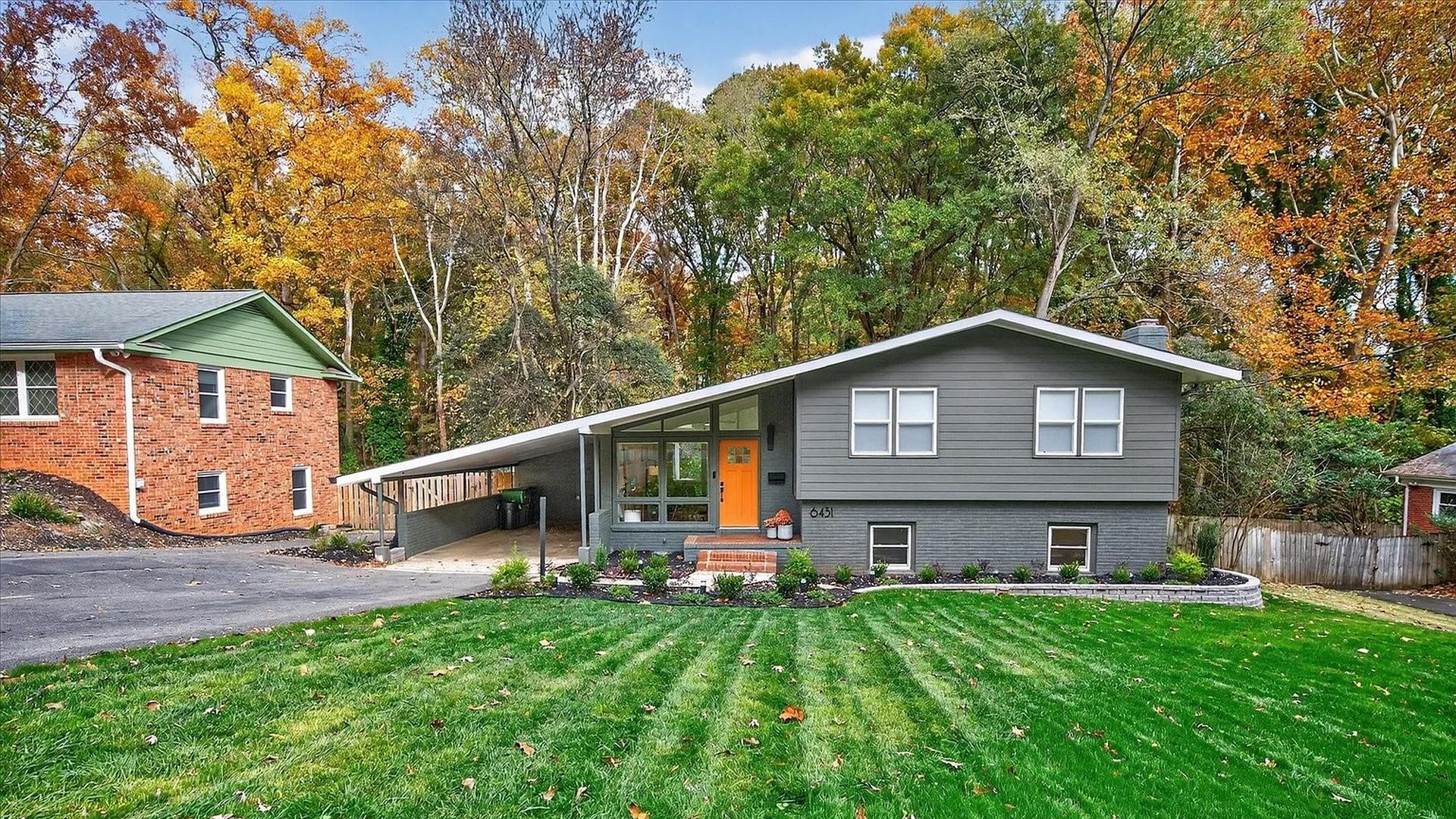 Gray mid-century modern house with an orange front door, large green lawn, and colorful autumn trees in the background under a clear blue sky.