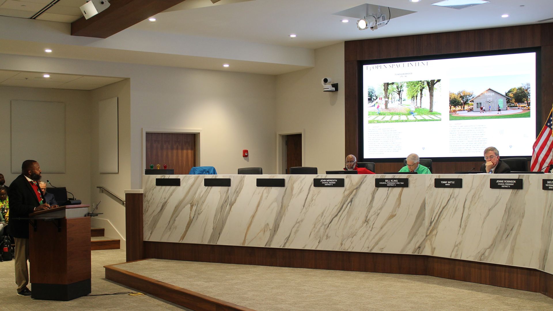 A man speaks at a wooden podium in a council chamber with five seated officials behind a marble-patterned desk, a large screen displaying images and text above them, and an American flag nearby.