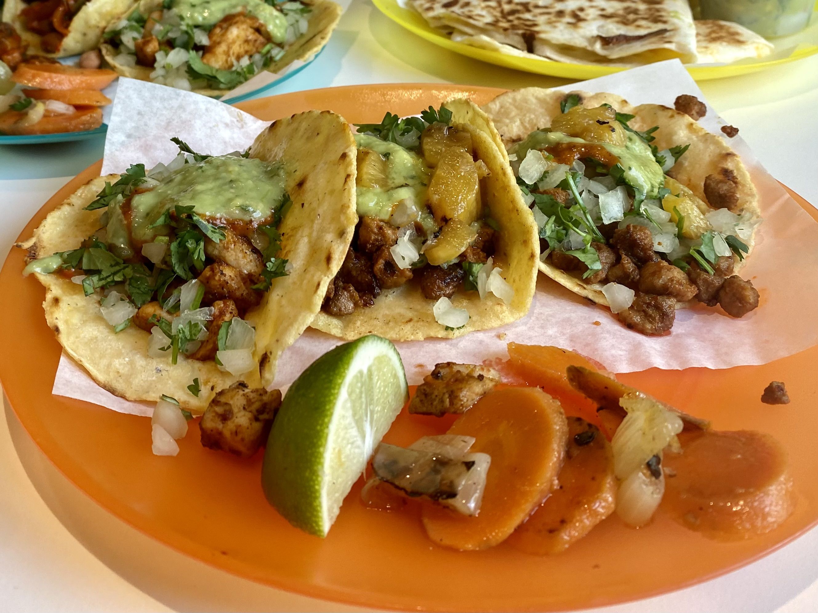 Three tacos with salsa verde on top with a slice of lime and carrots and onions on an orange plate.
