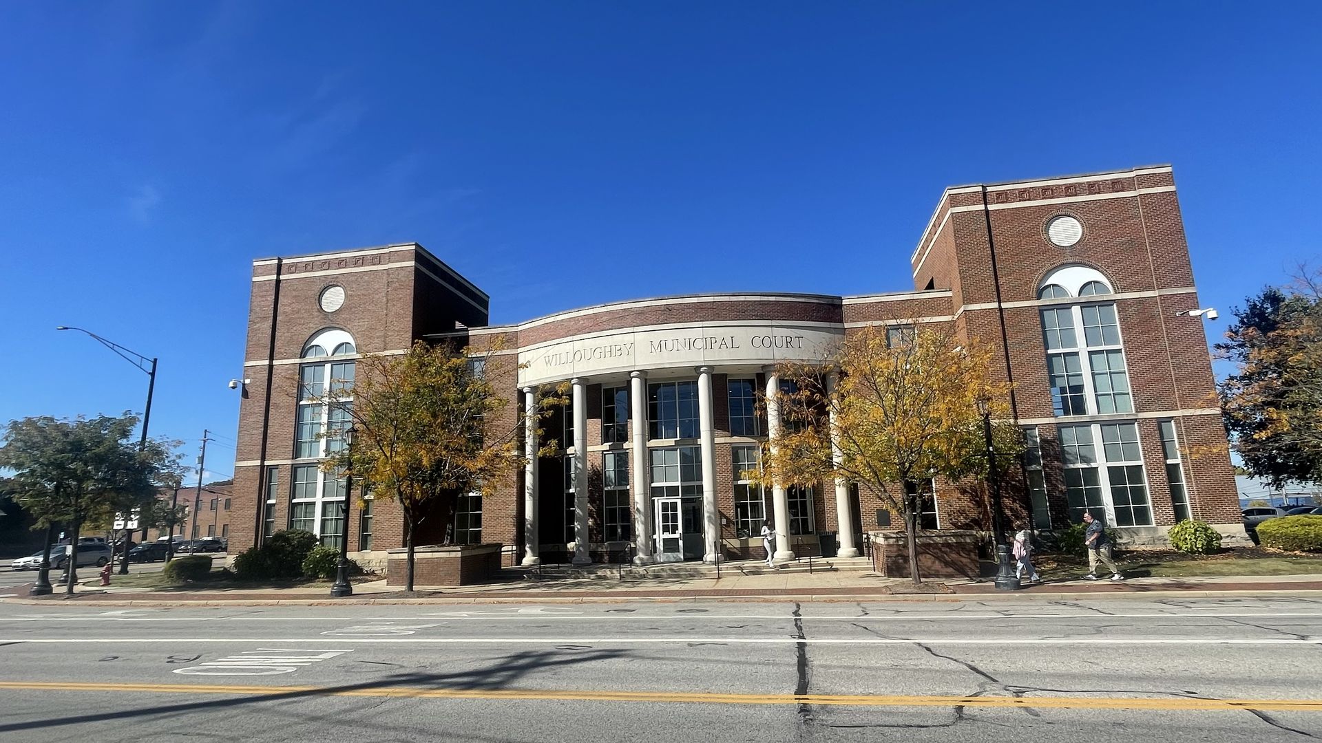 Willoughby Municipal Court building: brick with white trim, curved entry, two tower-like wings, arched windows, blue sky, autumn trees, and a street with pedestrians.