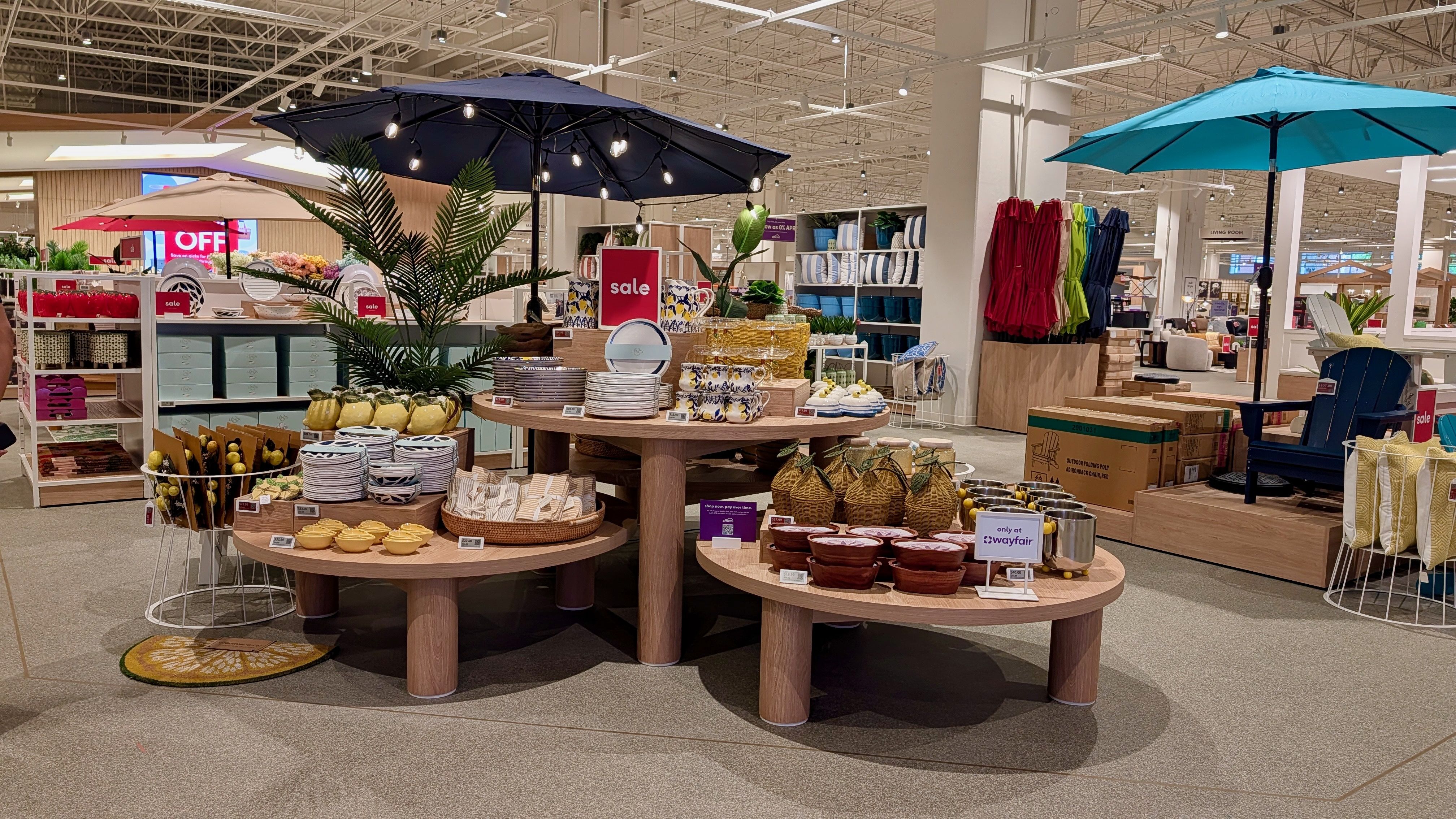 Inside a bright home goods store, circular wooden tables display plates, bowls, mugs, and baskets. A large plant, string lights, and blue/turquoise umbrellas surround the sale displays in the background.