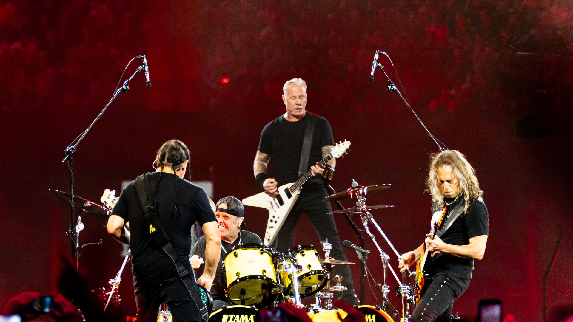 Rock band on a red-lit stage: a guitarist with a white Flying V center stage, two other players, a Tama drum kit in front, and a large crowd silhouetted in the background.