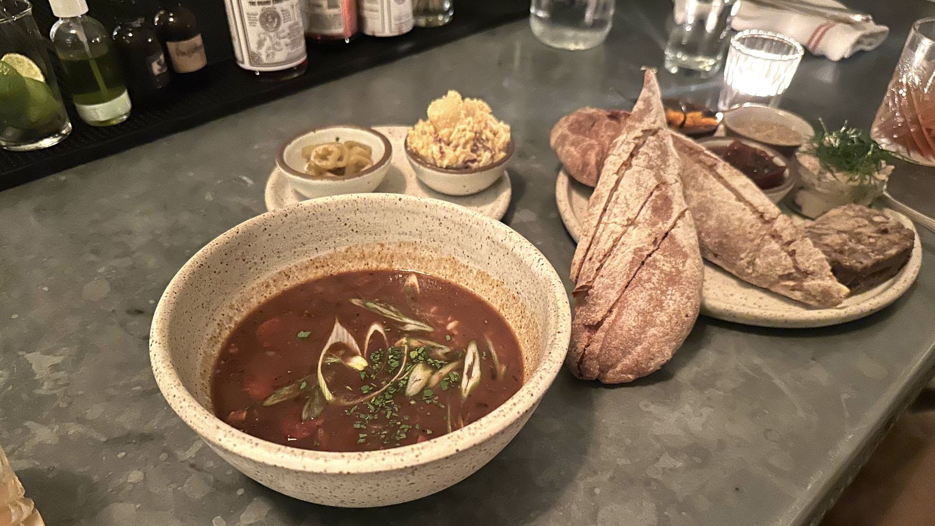 A bowl of gumbo sits on a countertop. Behind it is a plate with two small bowls of banana peppers and potato salad, and a plate of rillete served with bread.