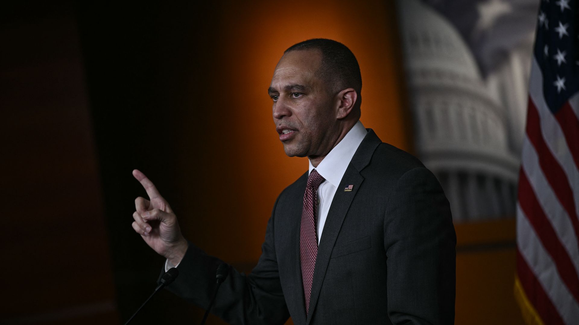 A man in a dark suit and red tie gestures with his right hand while speaking in front of microphones, with an American flag and a blurred government building backdrop behind him.