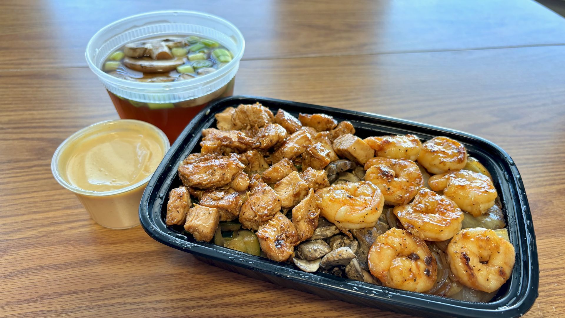 Black takeout container with grilled shrimp, chicken, mushrooms, and onions, served with a cup of dipping sauce and a clear cup of broth with mushrooms and green onions on a wooden table.