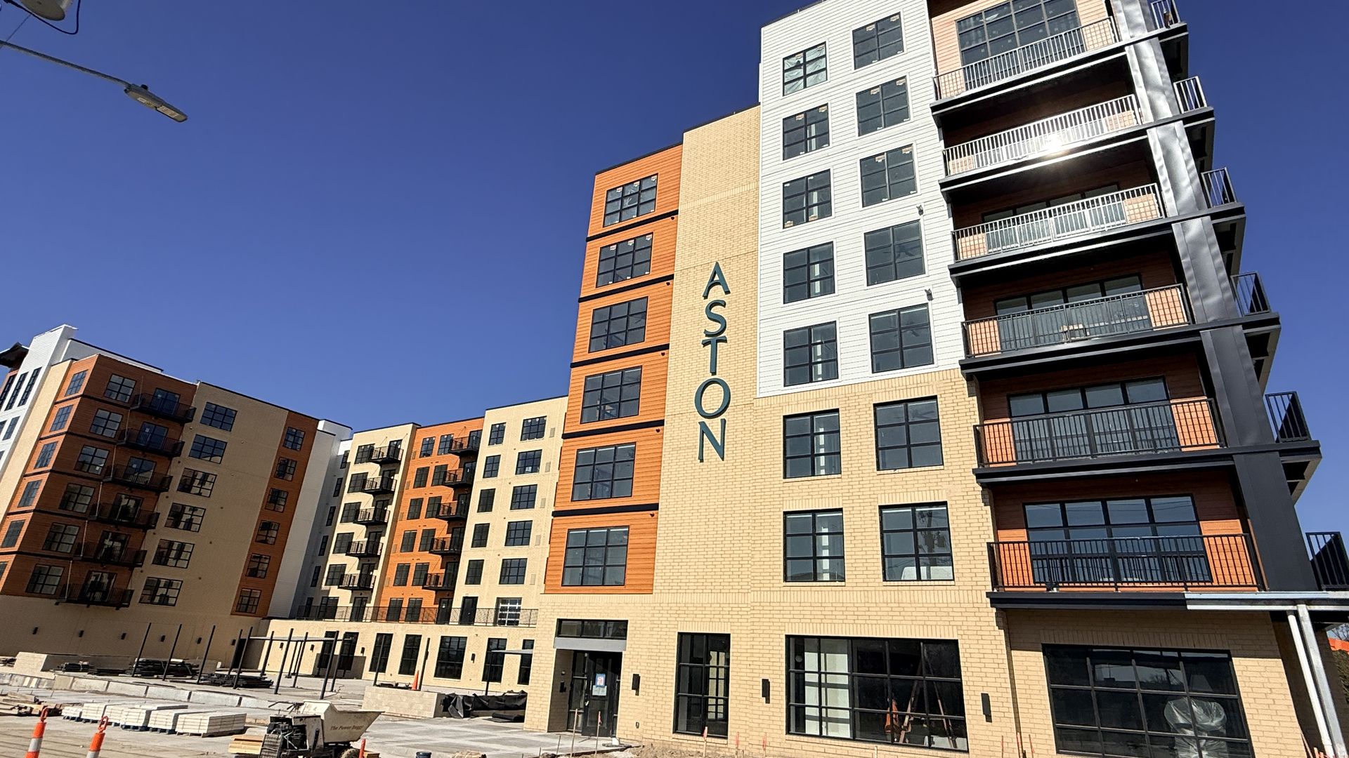 Under-construction modern apartment building with beige and orange brick, many windows and balconies on the right; construction equipment and orange cones in the foreground, blue sky.
