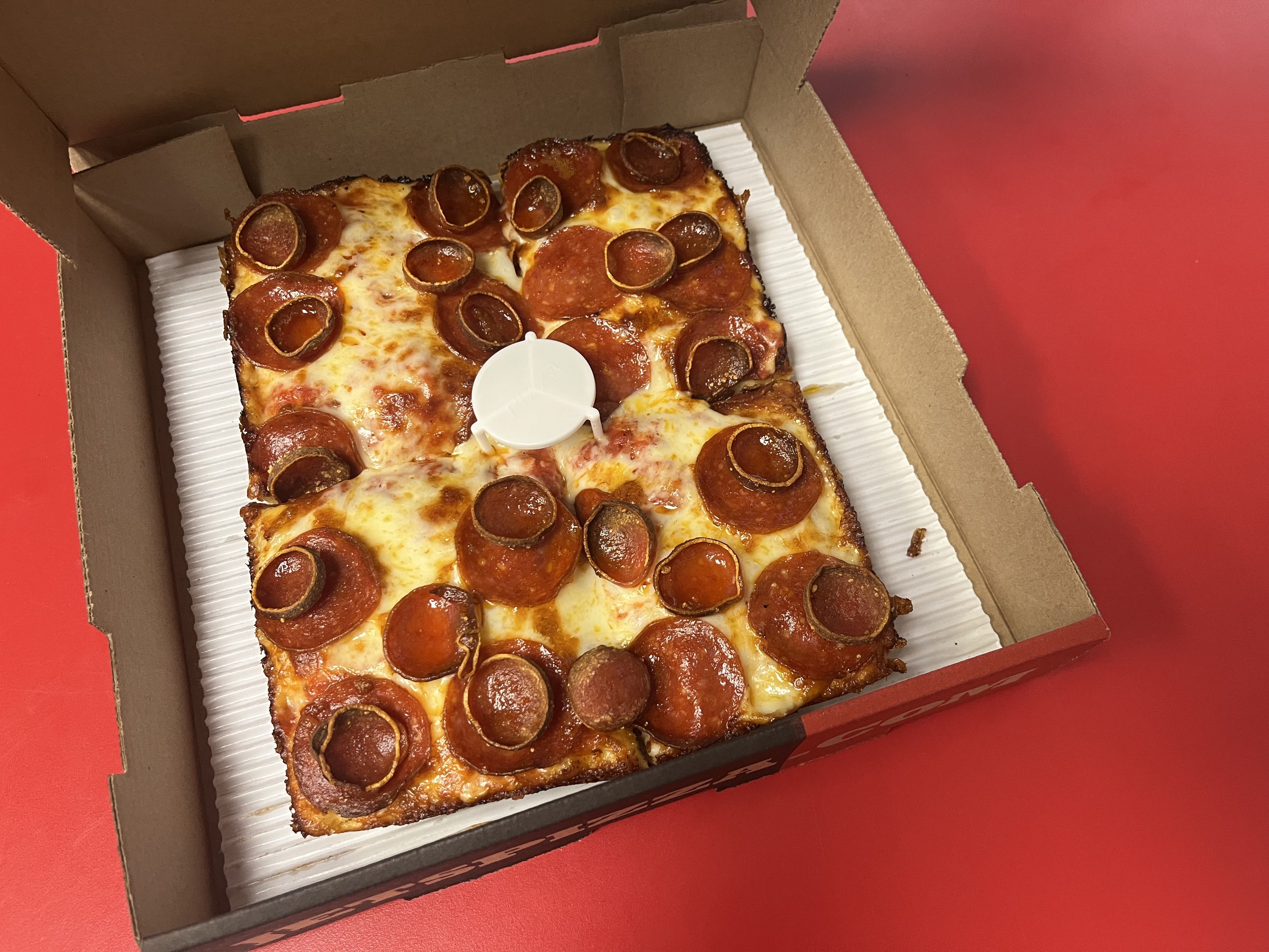 Open pizza box reveals a rectangular pepperoni pizza with curled pepperoni cups, melted cheese, and browned crust; a white plastic pizza saver sits in the center.