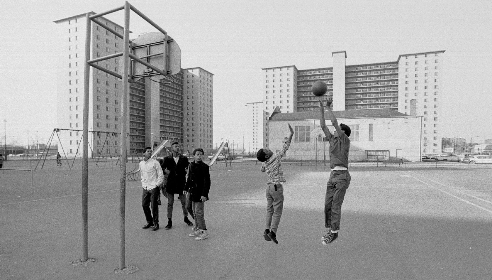 Photo of kids playing basketball in front of buildings while in their street clothes. 