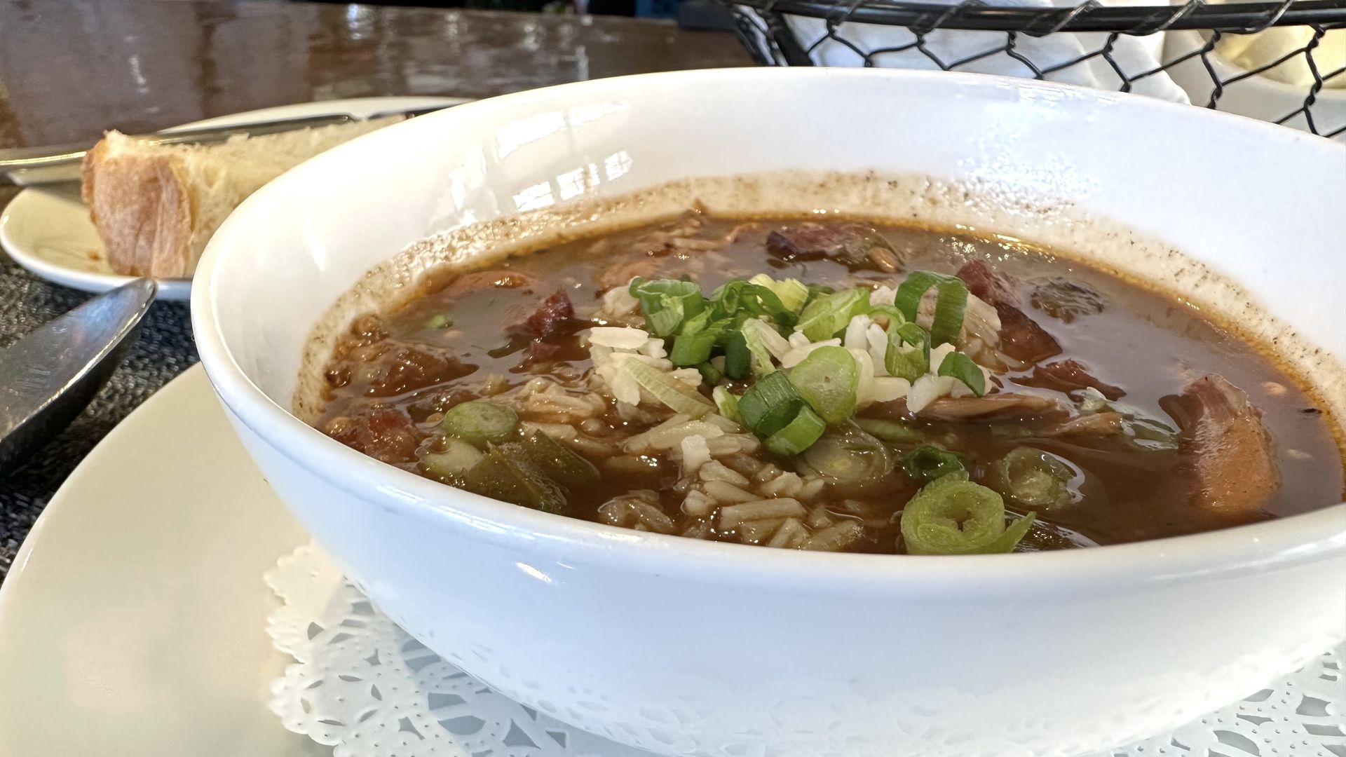 A white bowl is filled with a gumbo, with rice and green onions. The bowl sits on a charger with a doily, and a piece of bread is visible in the background.