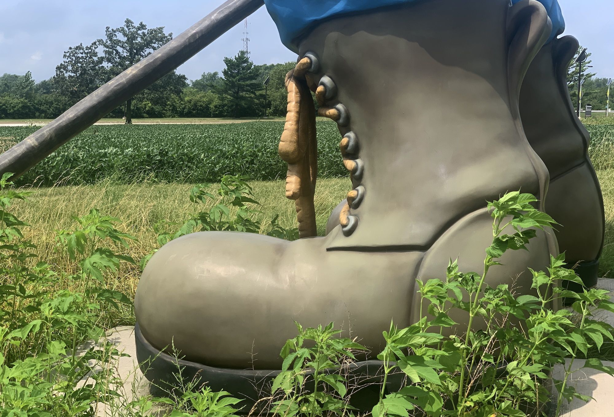 Close-up of a large, grayish-brown boot sculpture with laces and a wooden pole, surrounded by green plants and a field under a partly cloudy sky.