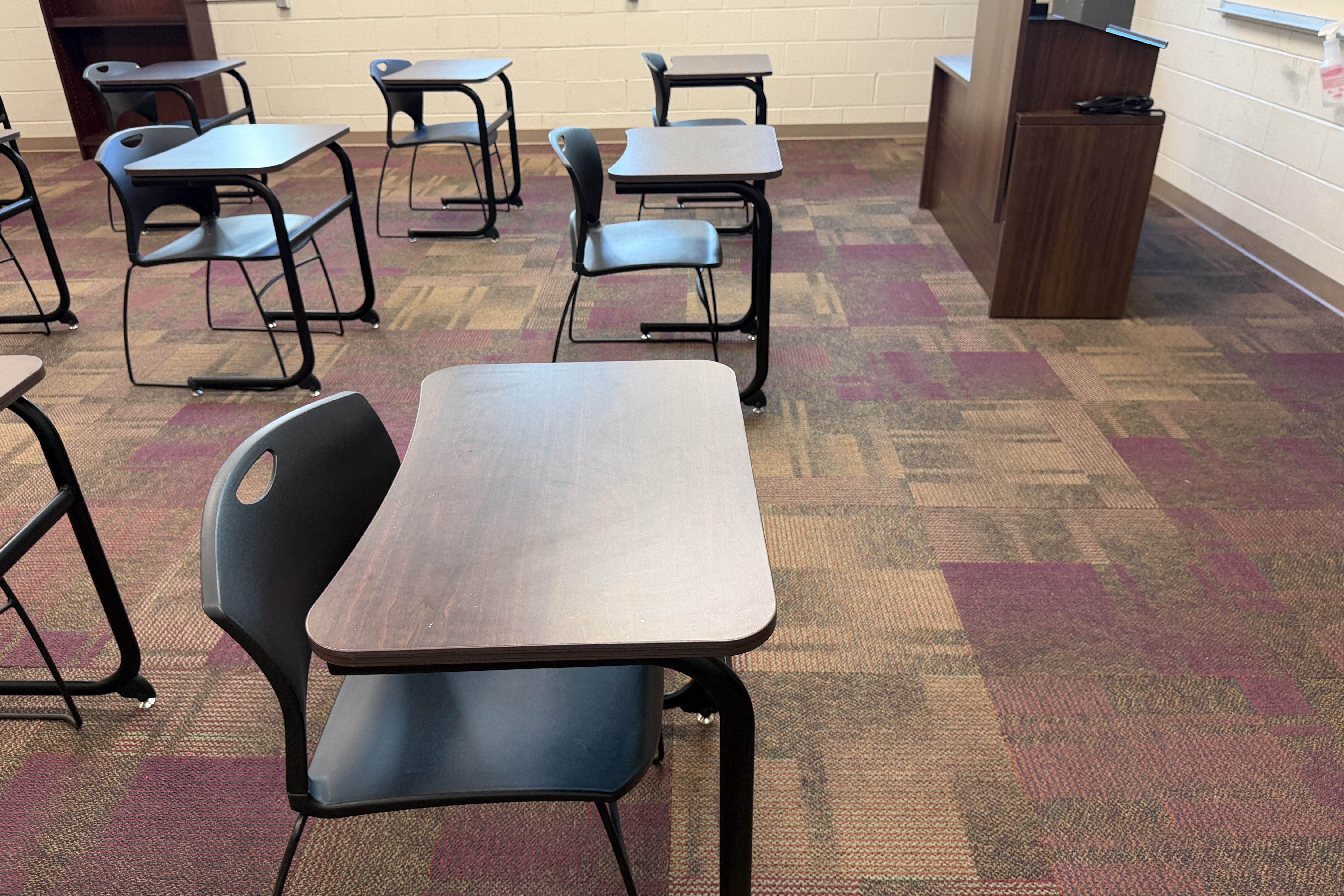 Empty classroom with rows of gray desks and black metal legs, chairs attached, a wooden podium at the front right, and a multicolor carpet in tan, maroon, and purple.