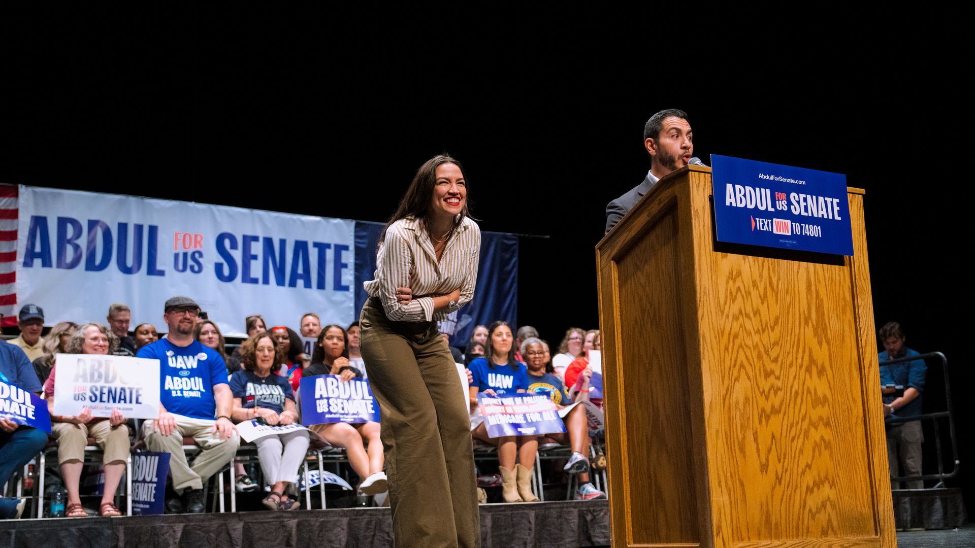 Alexandria Ocasio-Cortez laughs to a crowd while Senate candidate Abdul El-Sayed speaks into a microphone.