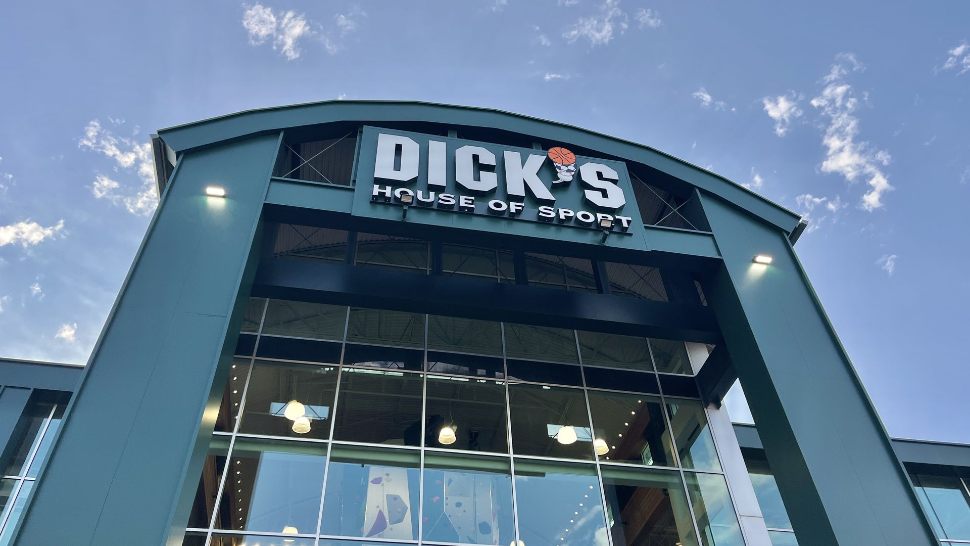 Exterior of a teal-colored Dick's House of Sport store with a large glass front and a sign featuring a basketball and sports shoe. Blue sky with scattered clouds above.