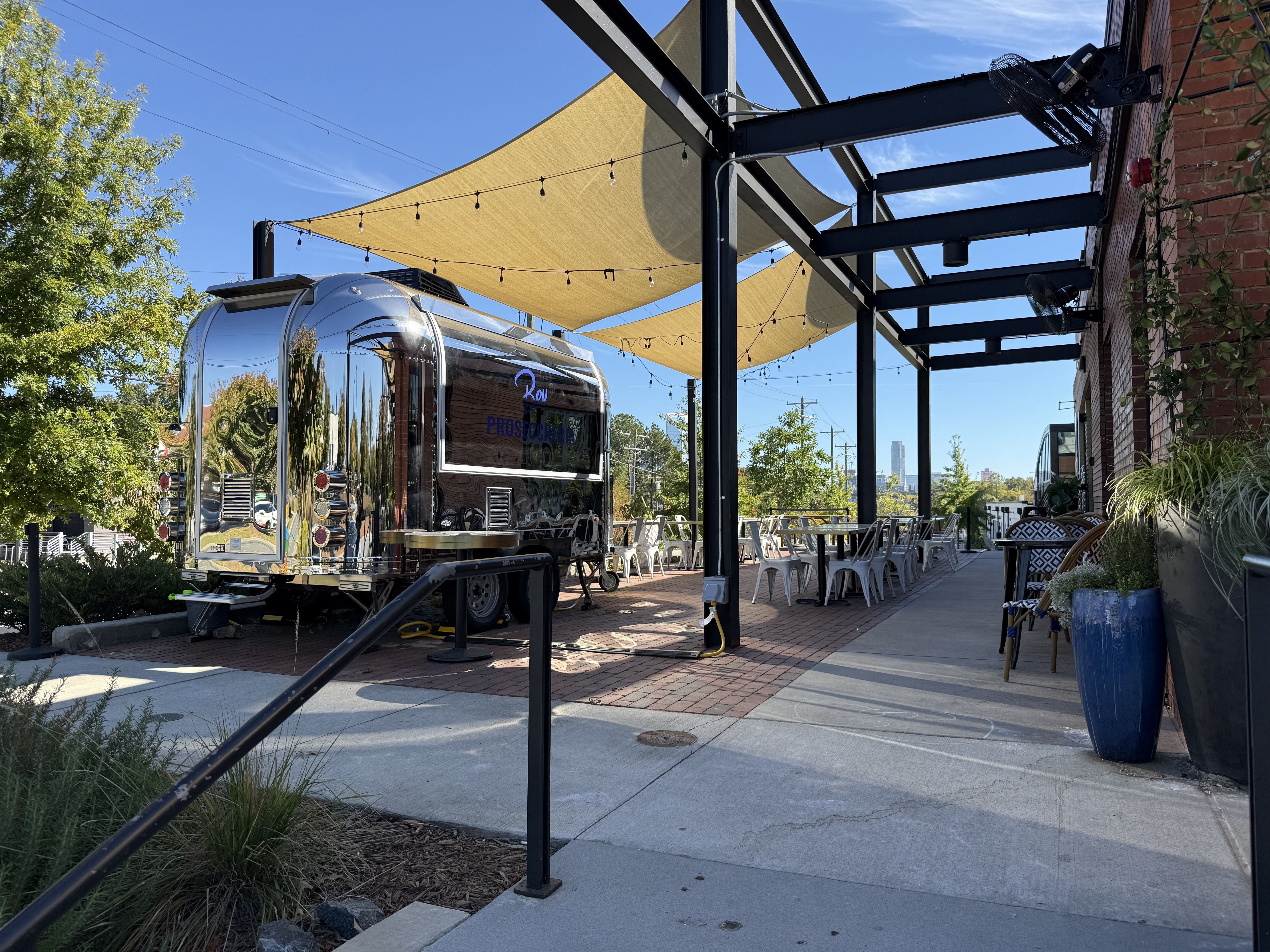 Shiny Airstream trailer in a brick-walled outdoor patio, under beige shade sails and string lights. Metal pergola beams, white chairs, potted plants, and a clear blue sky.